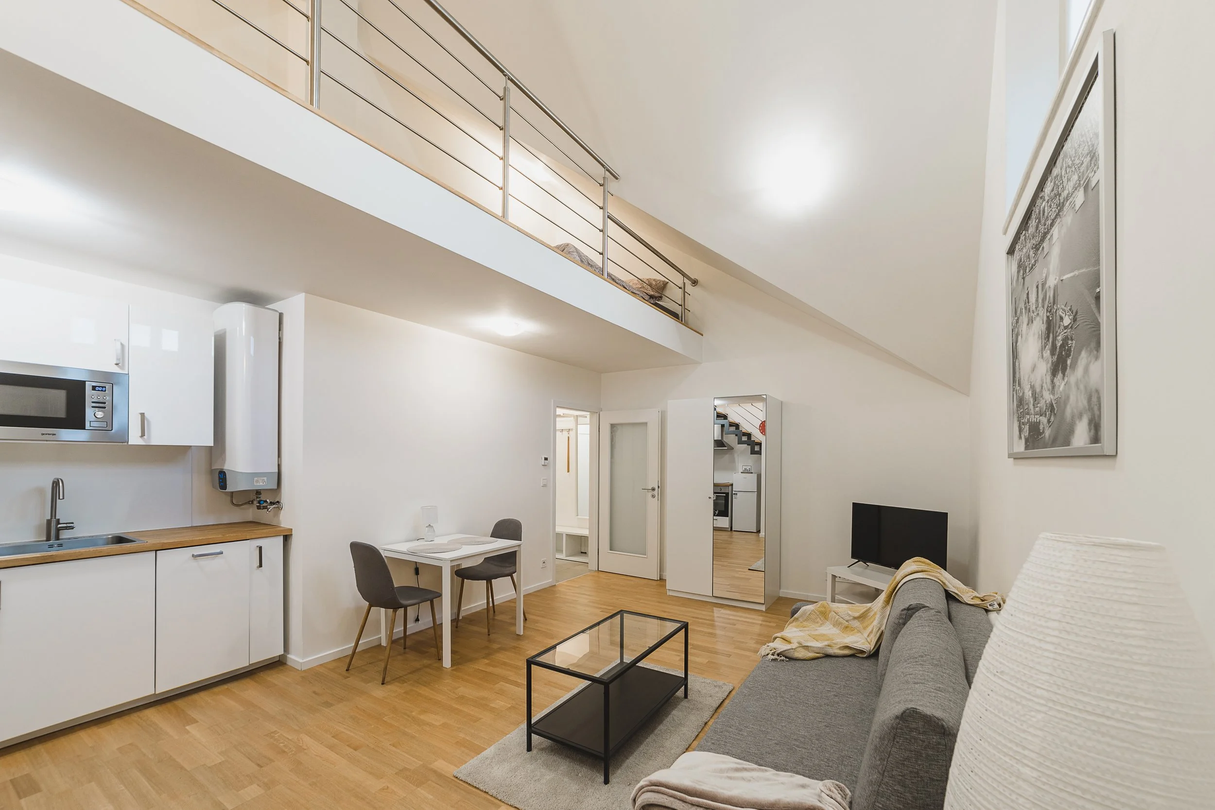 Living room with gray sofa, white armchair, glass coffee table, small dining table with two chairs, kitchen area with white cabinets and microwave, mounted water heater, and artwork on the wall. Loft area with a bed and railing above the living space