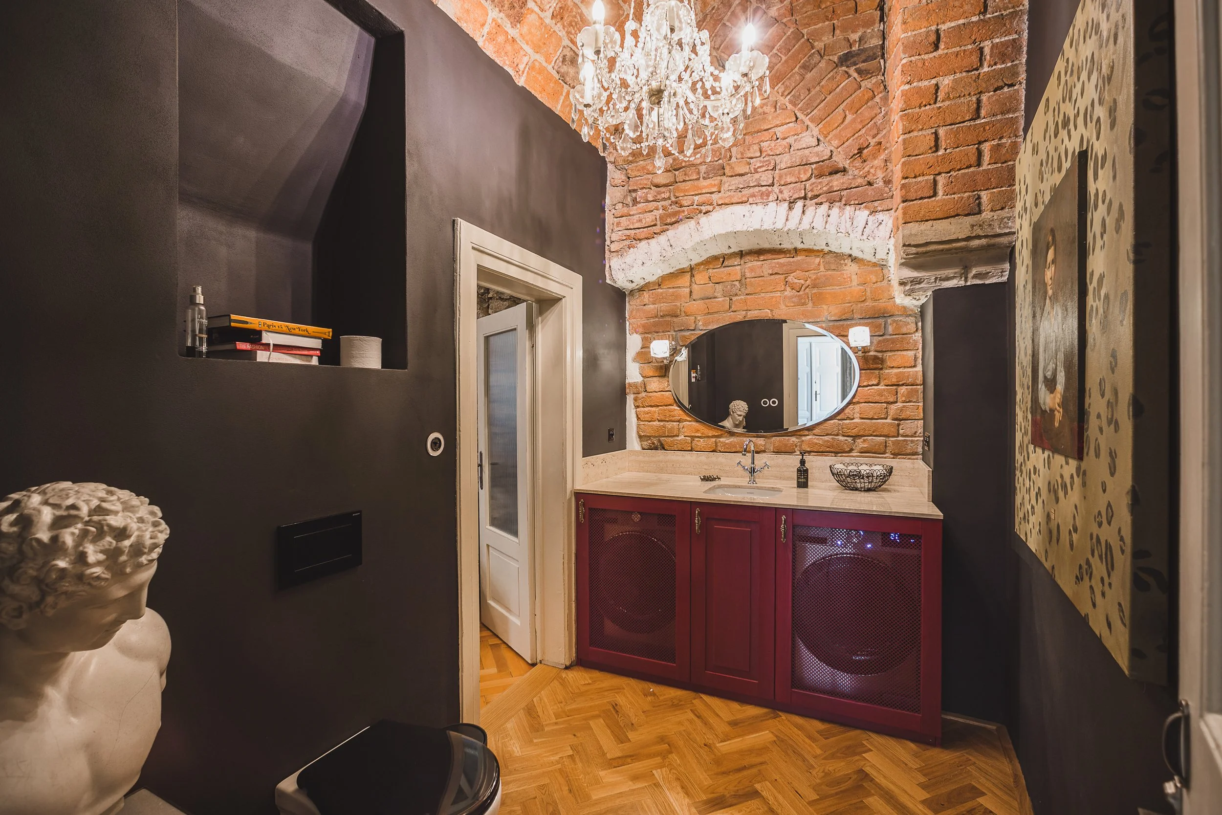 Interior of a stylish room with exposed brick walls, black painted walls, a chandelier, a vanity with red cabinet doors and a beige countertop, an oval mirror, and decorative items including books, a bust sculpture, and a bowl.