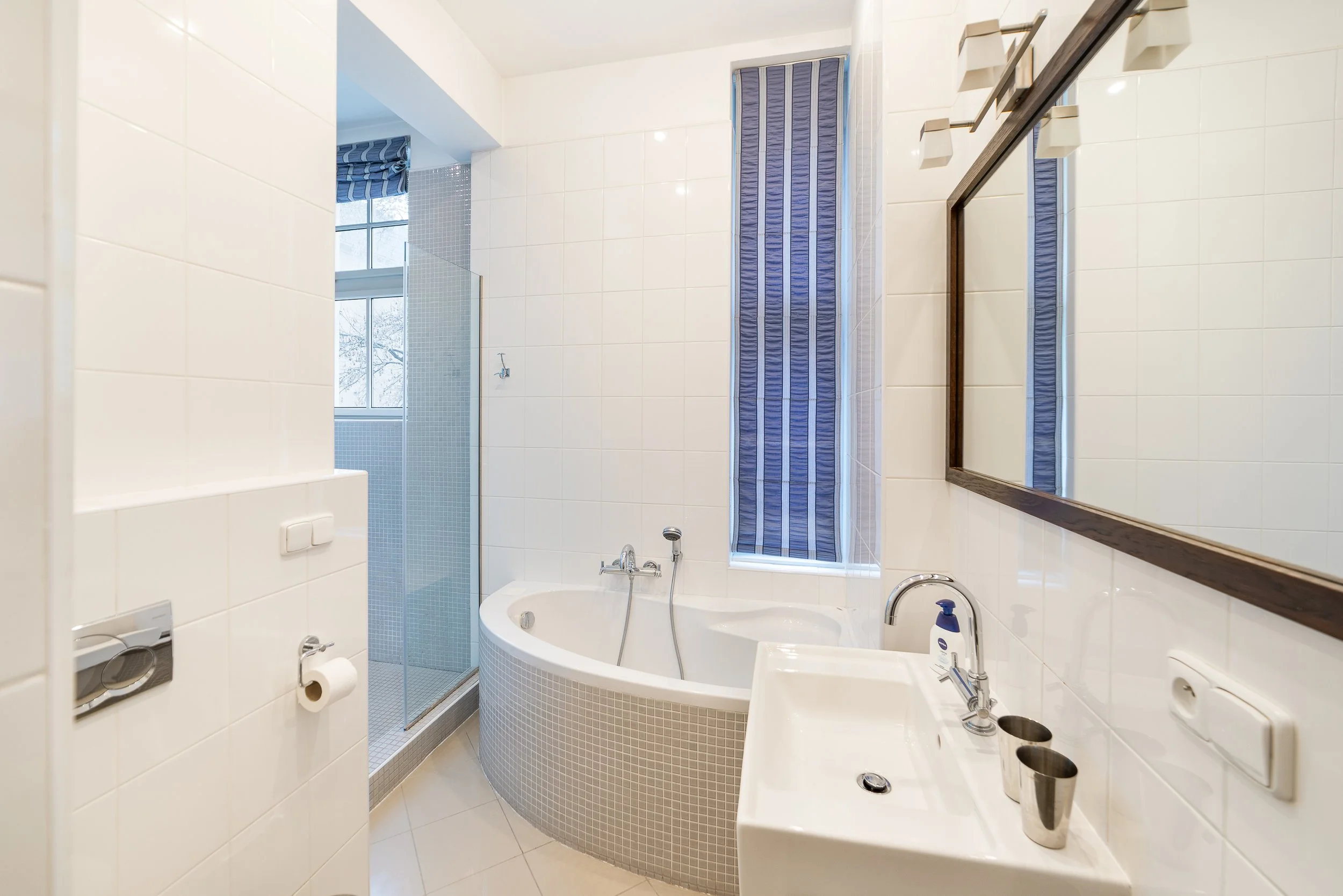 Bright bathroom with a corner bathtub, a glass-enclosed shower, a large mirror with a wooden frame, and a window with blue-striped curtains.