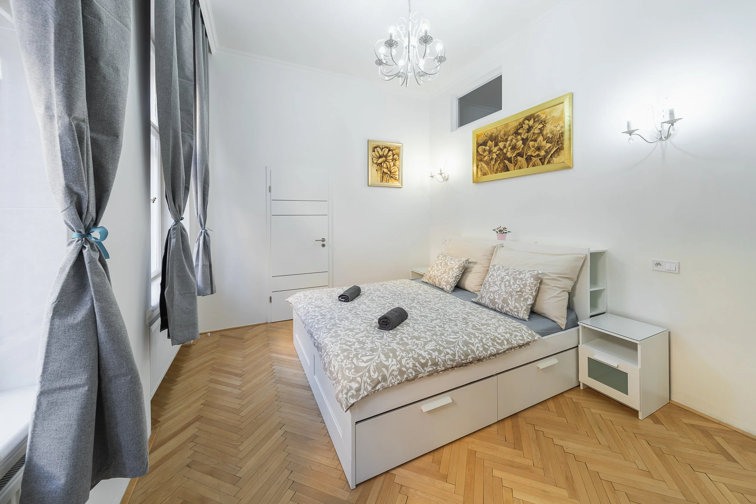 A bright bedroom with a white bed and floral bedding, two rolled towels, gray curtains, wall art with floral themes, parquet wood flooring, and a decorative chandelier.