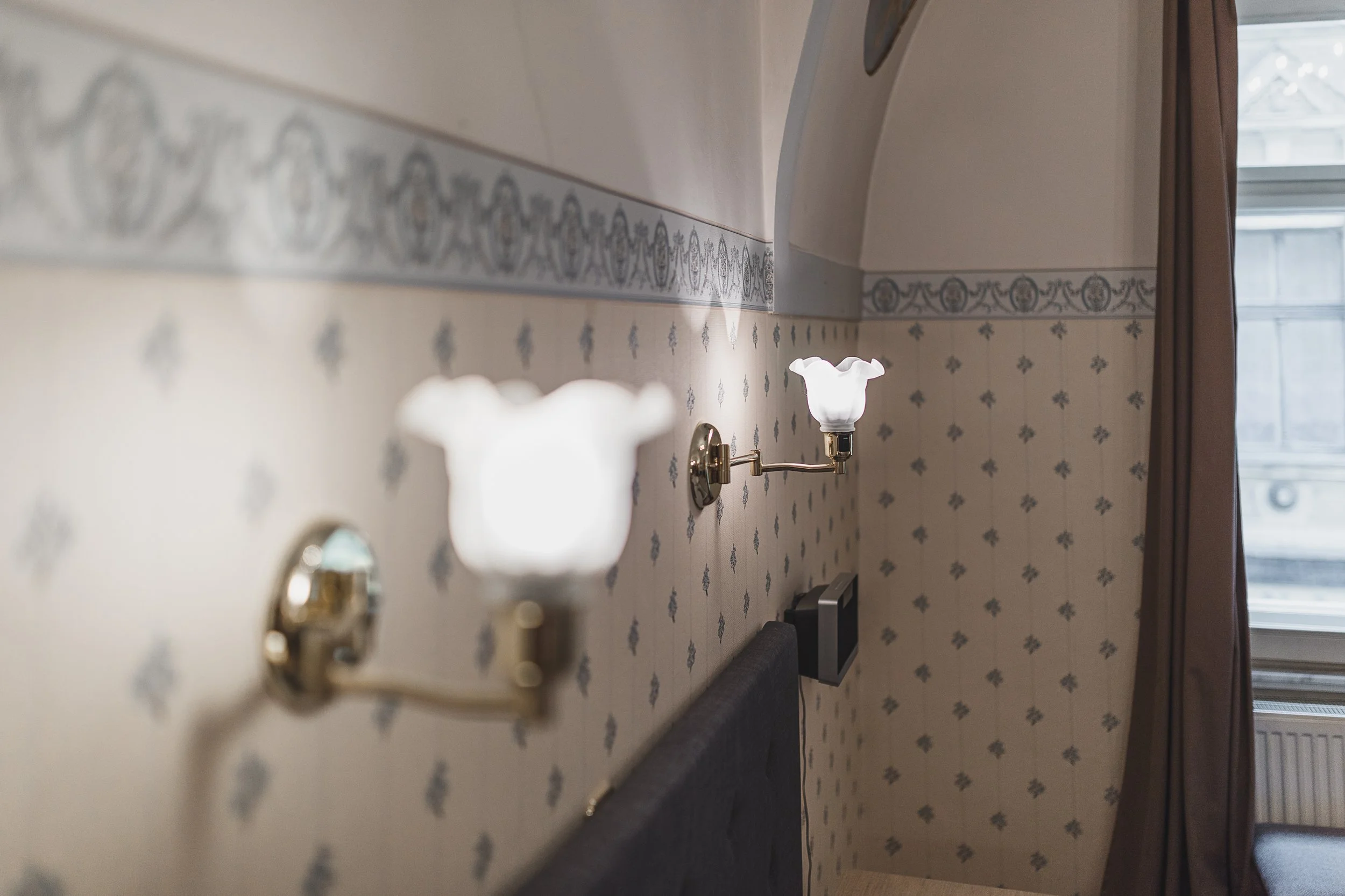 Interior of a room with floral-patterned wallpaper, featuring a wall-mounted light fixture with a decorative lampshade near a window with brown curtains.