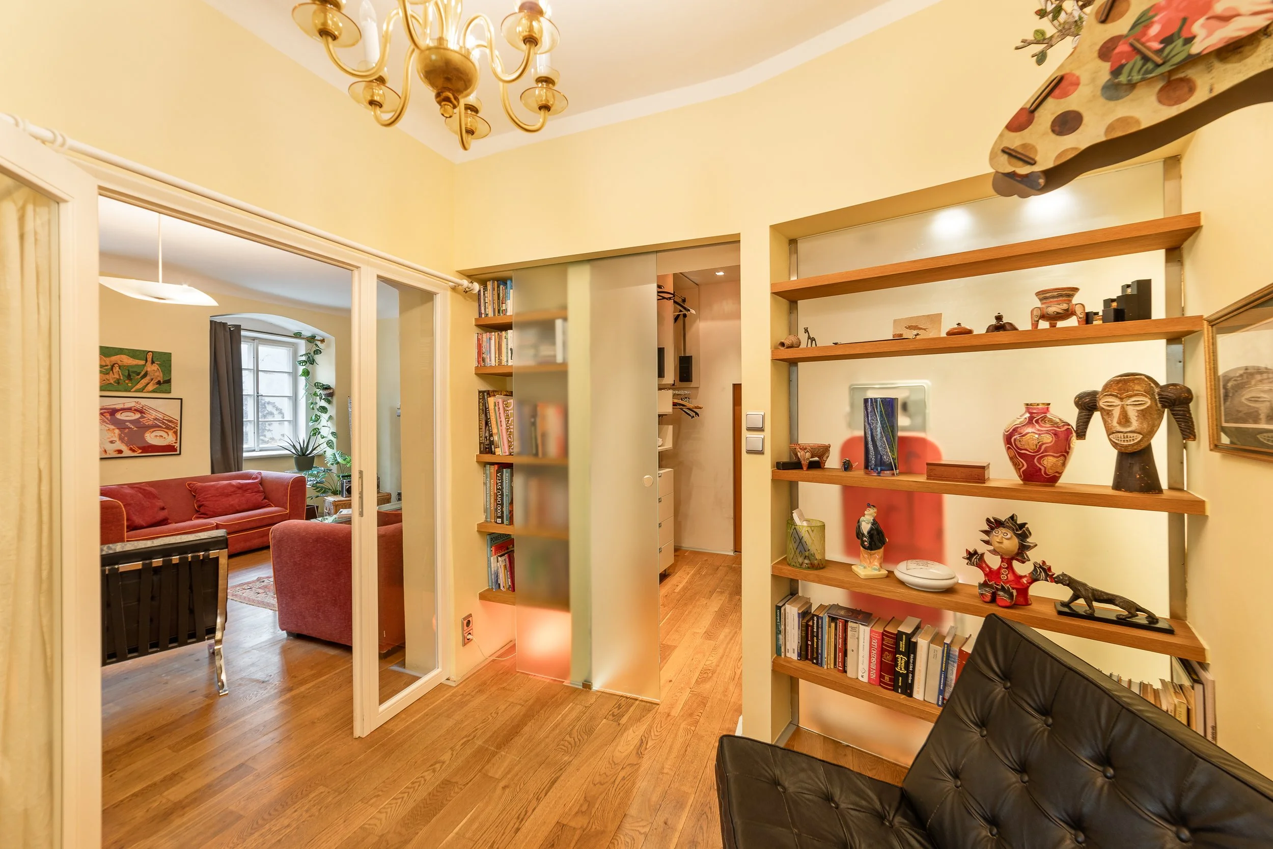 Interior of a cozy living room with wooden floors, yellow walls, a black leather chair, and open shelves displaying decorative items and books. There is a dining area visible in the background with a red couch, a window with curtains, and wall art. A