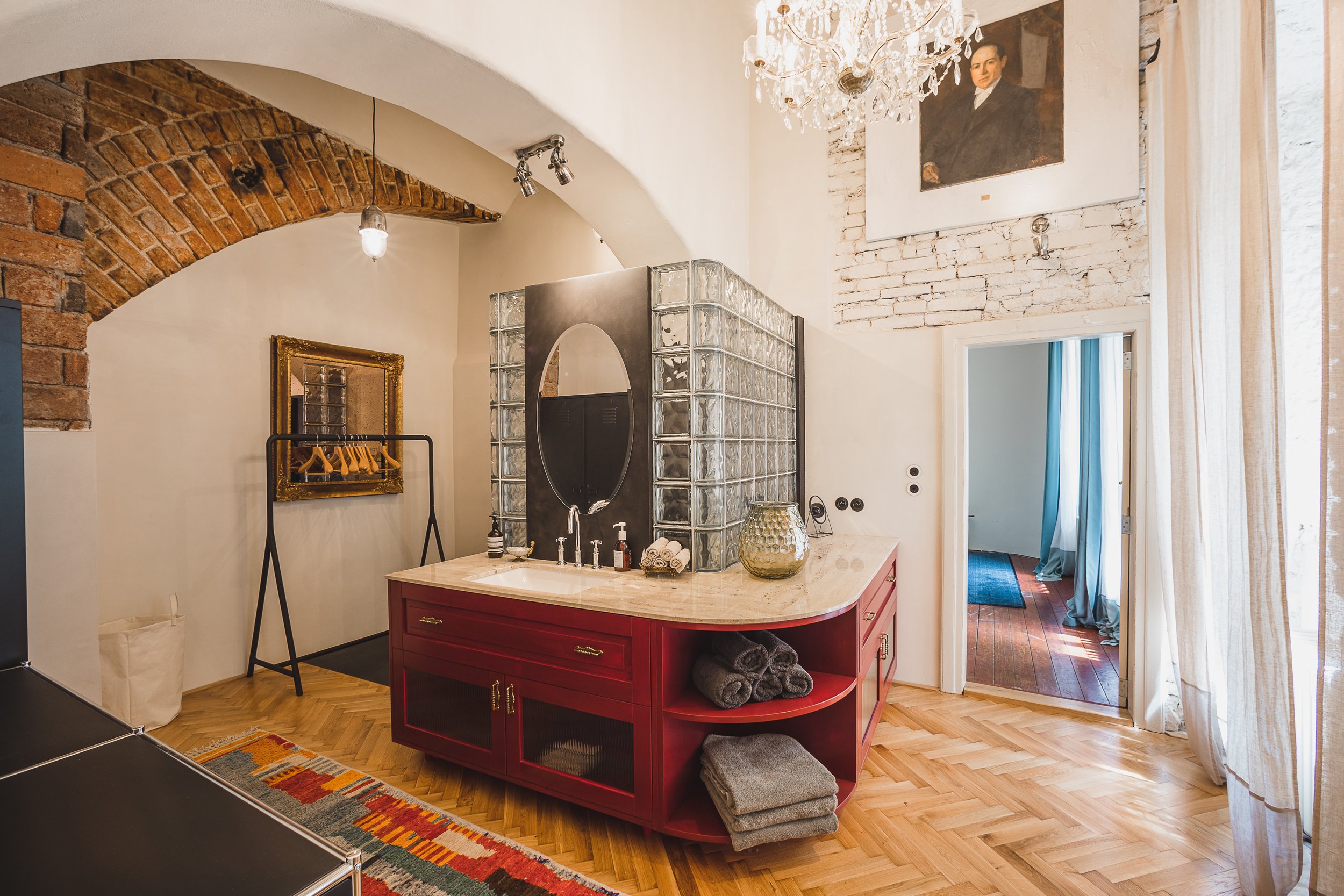 Interior of a room with brick archway and a chandelier, with a red kitchen island, glass block wall, framed mirror, and artwork on the white brick wall.