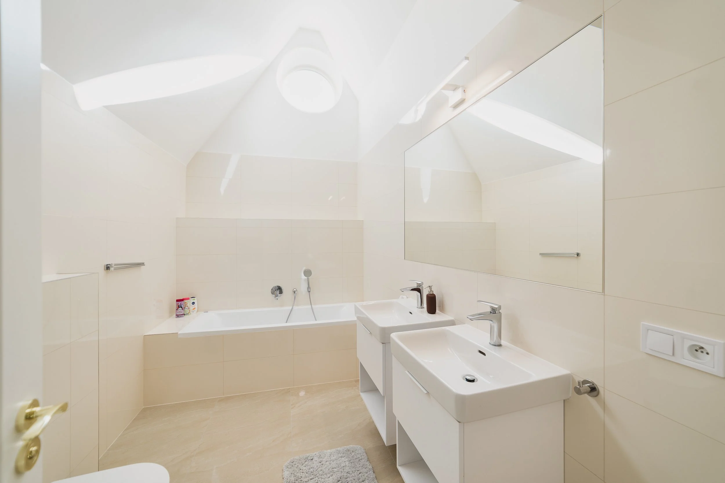 Modern bathroom with beige tiled walls and floor, featuring a bathtub, double sinks, a large mirror, and minimal decor.