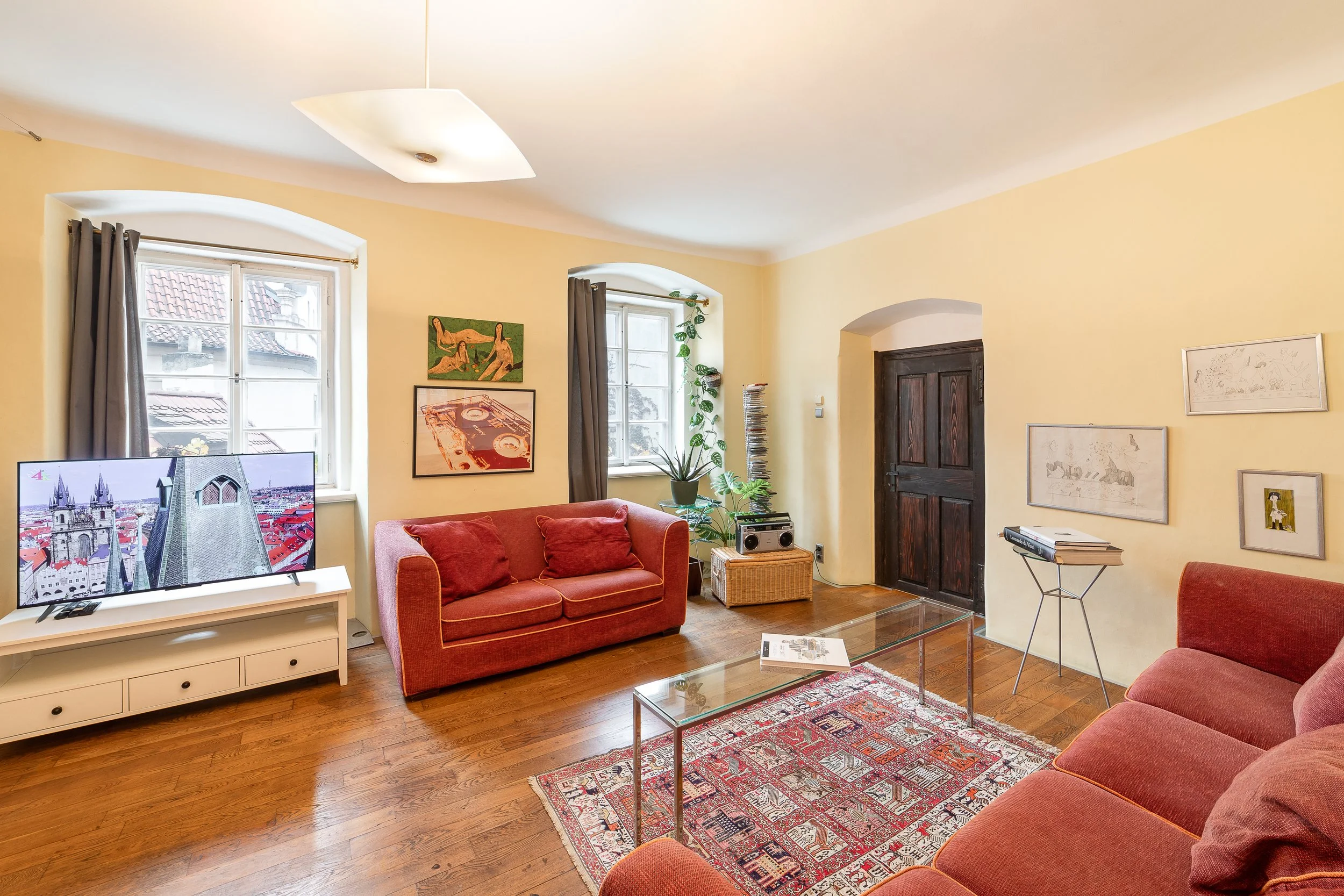 Living room with yellow walls, wooden floor, two windows with gray curtains, a red sofa with cushions, a glass coffee table, artwork on the walls, a flat-screen TV, and a potted plant near the window.