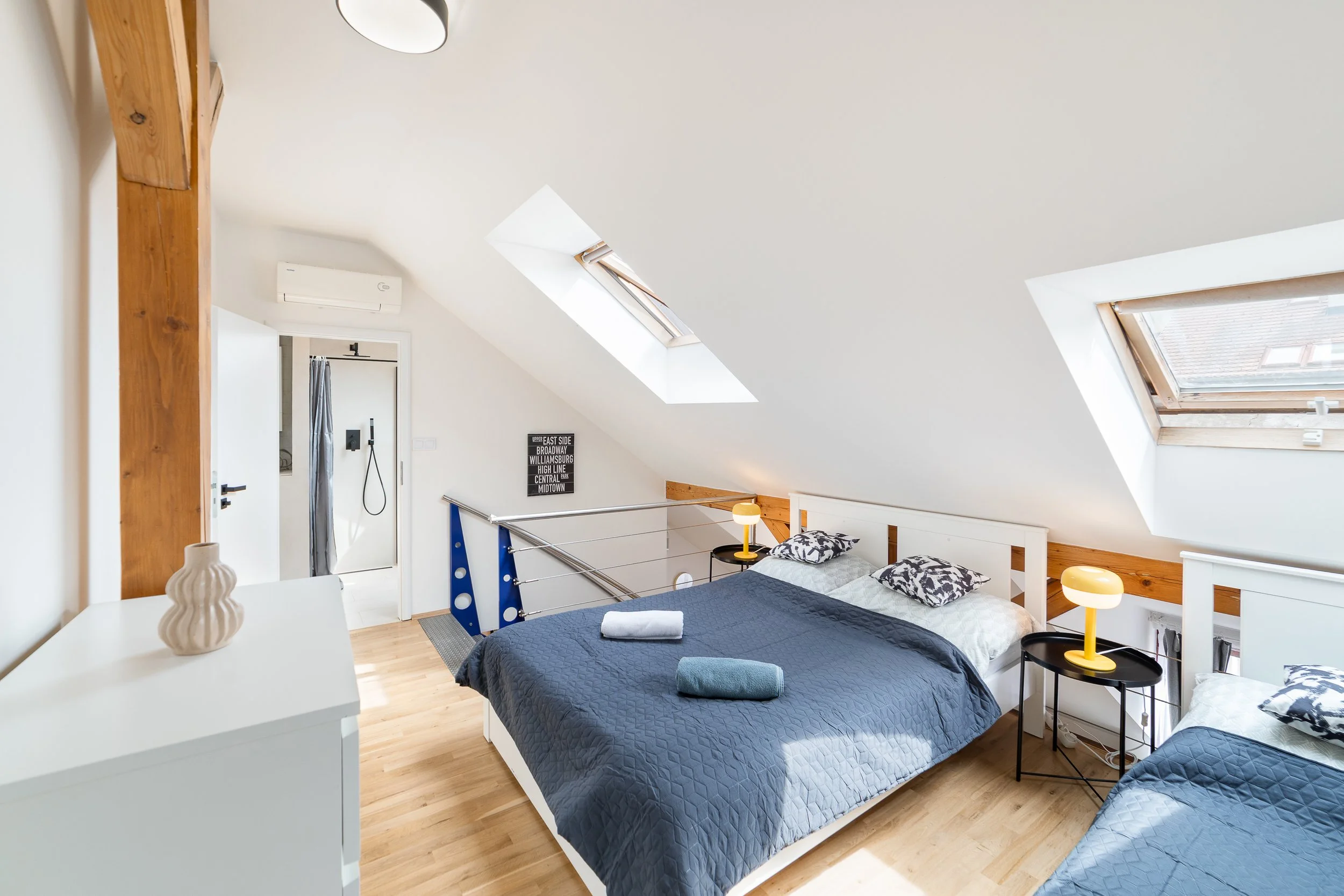 A bright attic bedroom with two beds, skylight windows, a wooden beam, and a small side table with a yellow lamp.