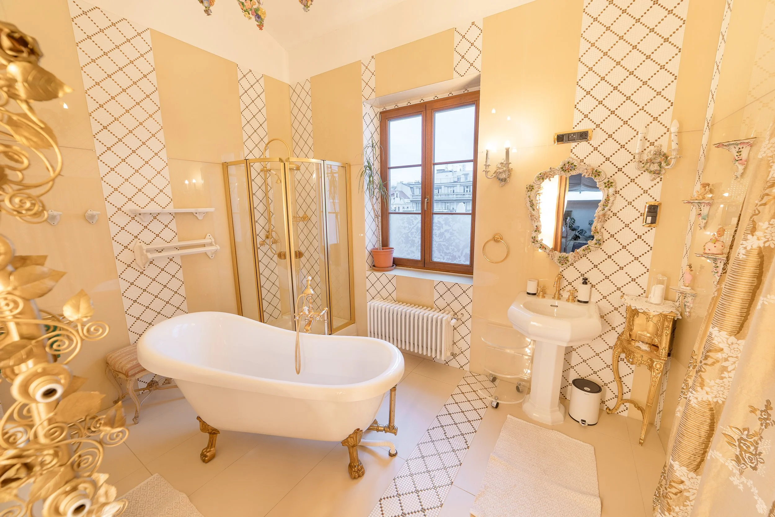 A luxurious bathroom with a vintage style, featuring a freestanding bathtub with ornate gold clawfoot legs, a separate glass shower stall, a window with wooden framing, and decorative accents such as floral wallpaper and gold fixtures.