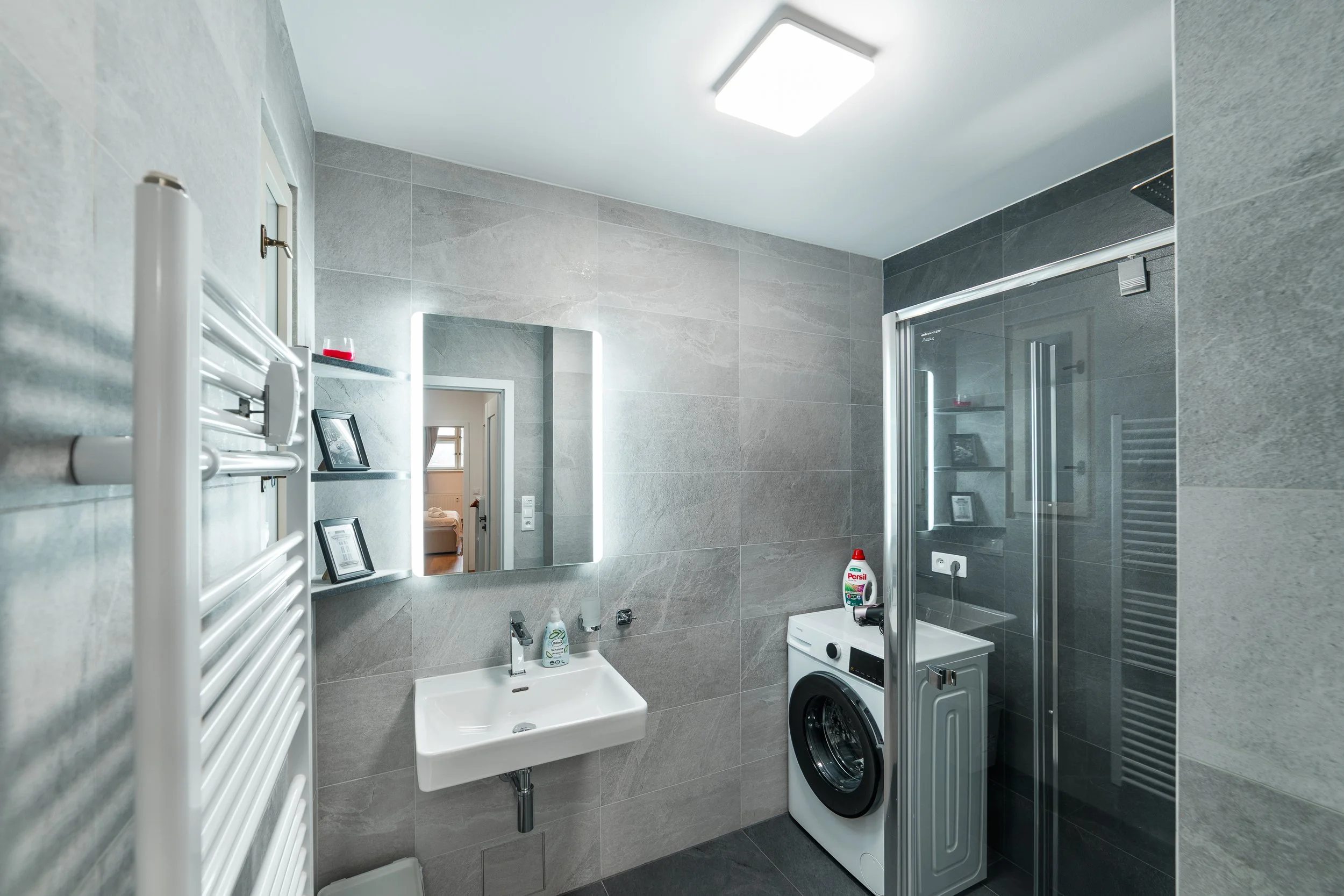 A modern bathroom with large gray tiles, a rectangular mirror with LED lighting, a white sink, a washing machine, and a shower with a glass door.