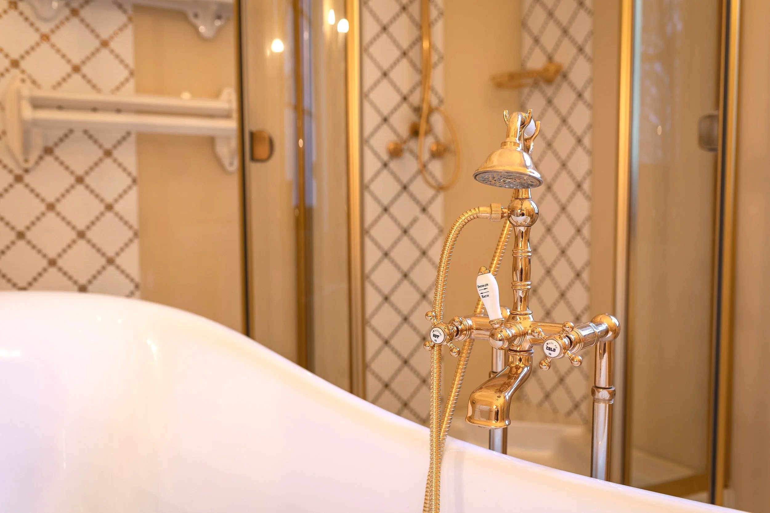 Close-up of a gold-toned vintage-style bathroom faucet with multiple handles labeled for different water temperatures, with a white bathtub in the foreground and a background featuring a shower with gold fixtures and patterned tiles.