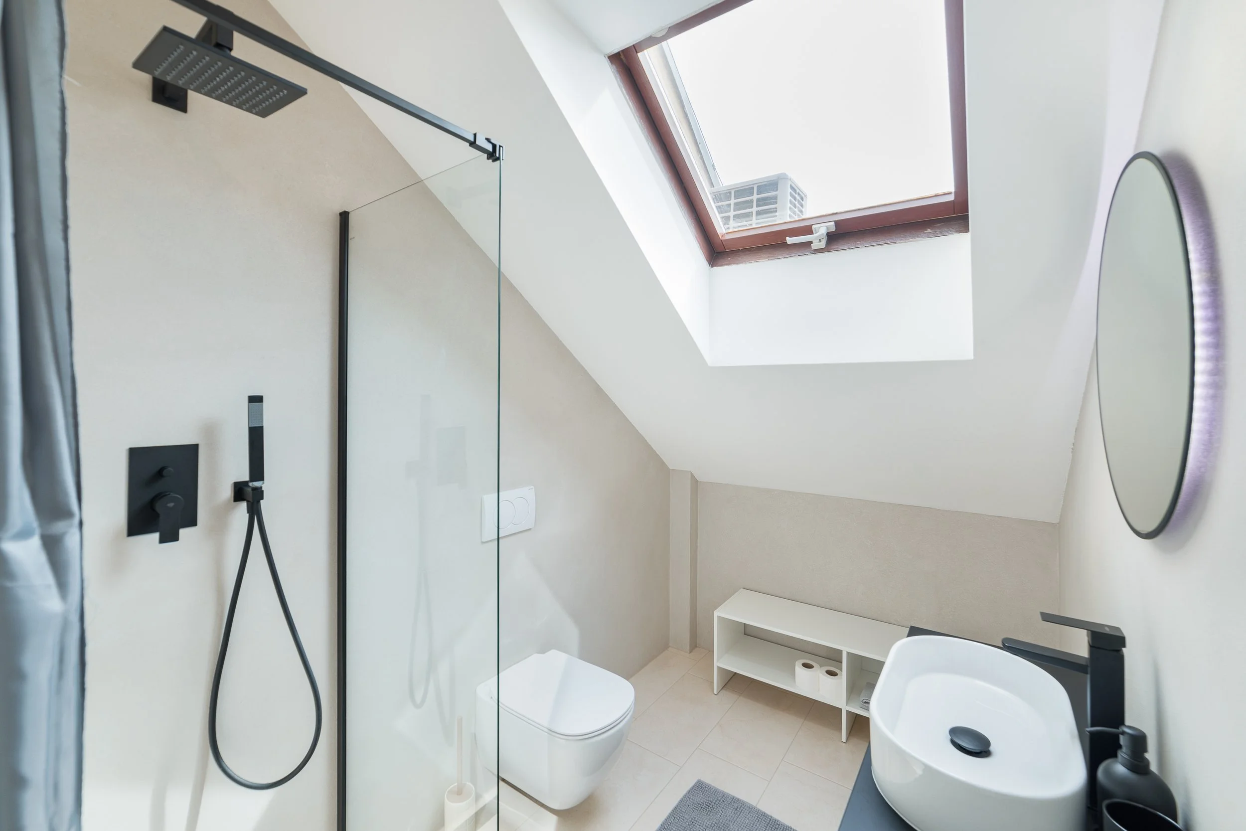 A contemporary bathroom with a skylight window, a walk-in shower with a rainfall showerhead, a wall-mounted toilet, a small white shelf, a round mirror, and a white sink with a black faucet.