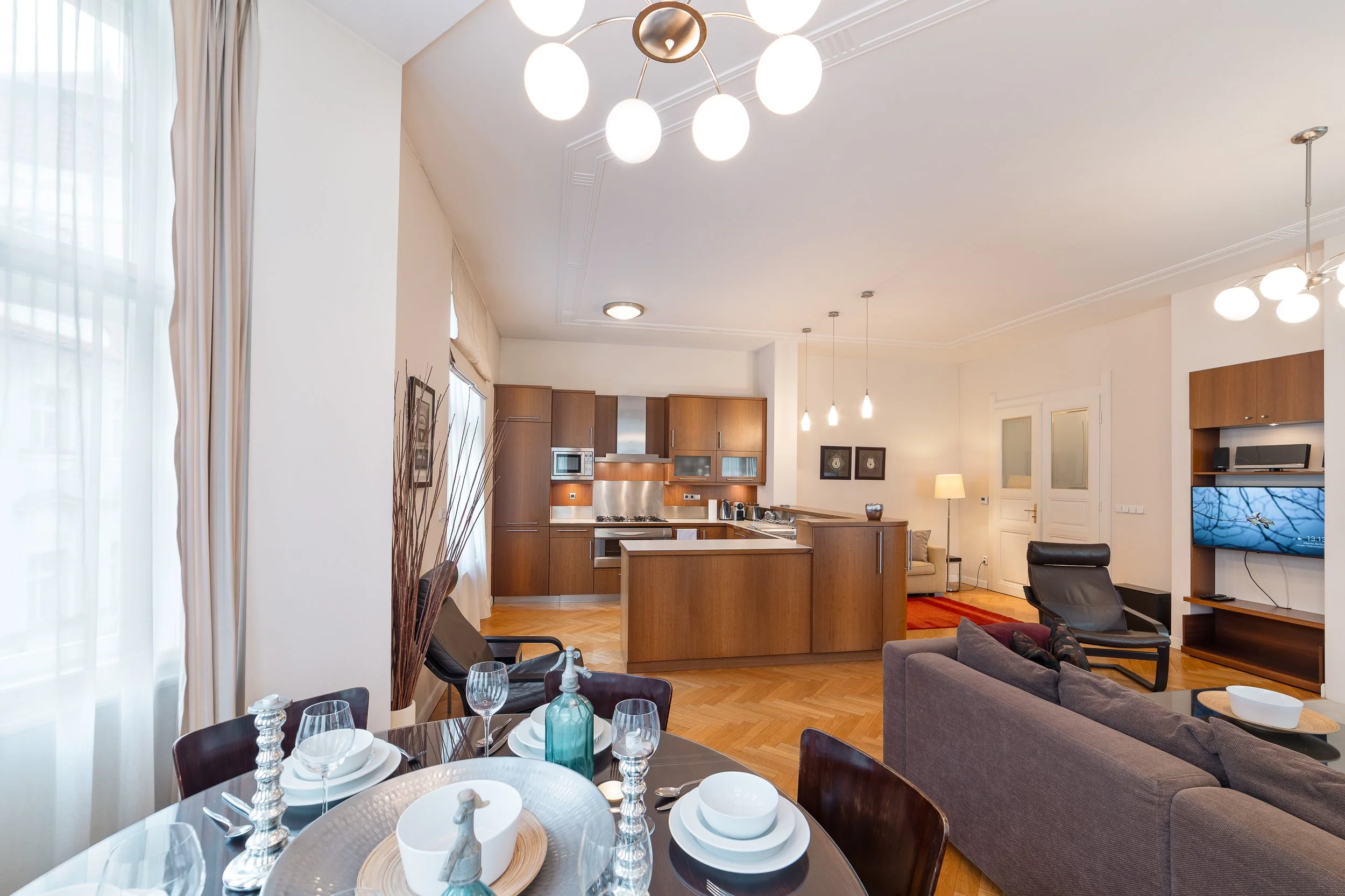 Open-concept living and dining area with wooden flooring, modern kitchen, gray sofa, TV, and a dining table set with white dishes and glassware.