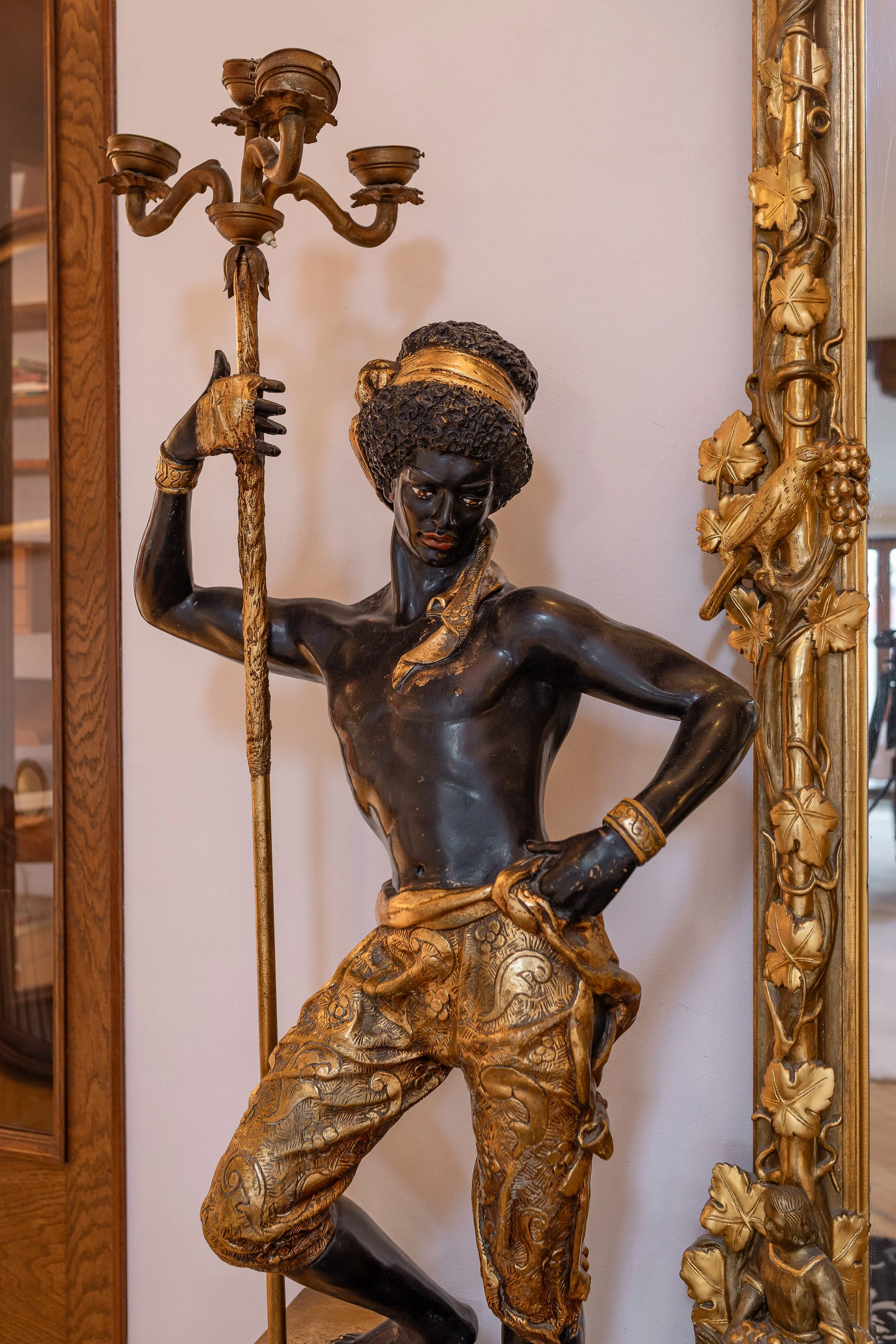 A detailed bronze sculpture of a woman with dark skin and curly hair, dressed in traditional clothing with gold accents, holding a staff and striking a dynamic pose, with a highly decorative gold frame with leaves and grapes beside her.