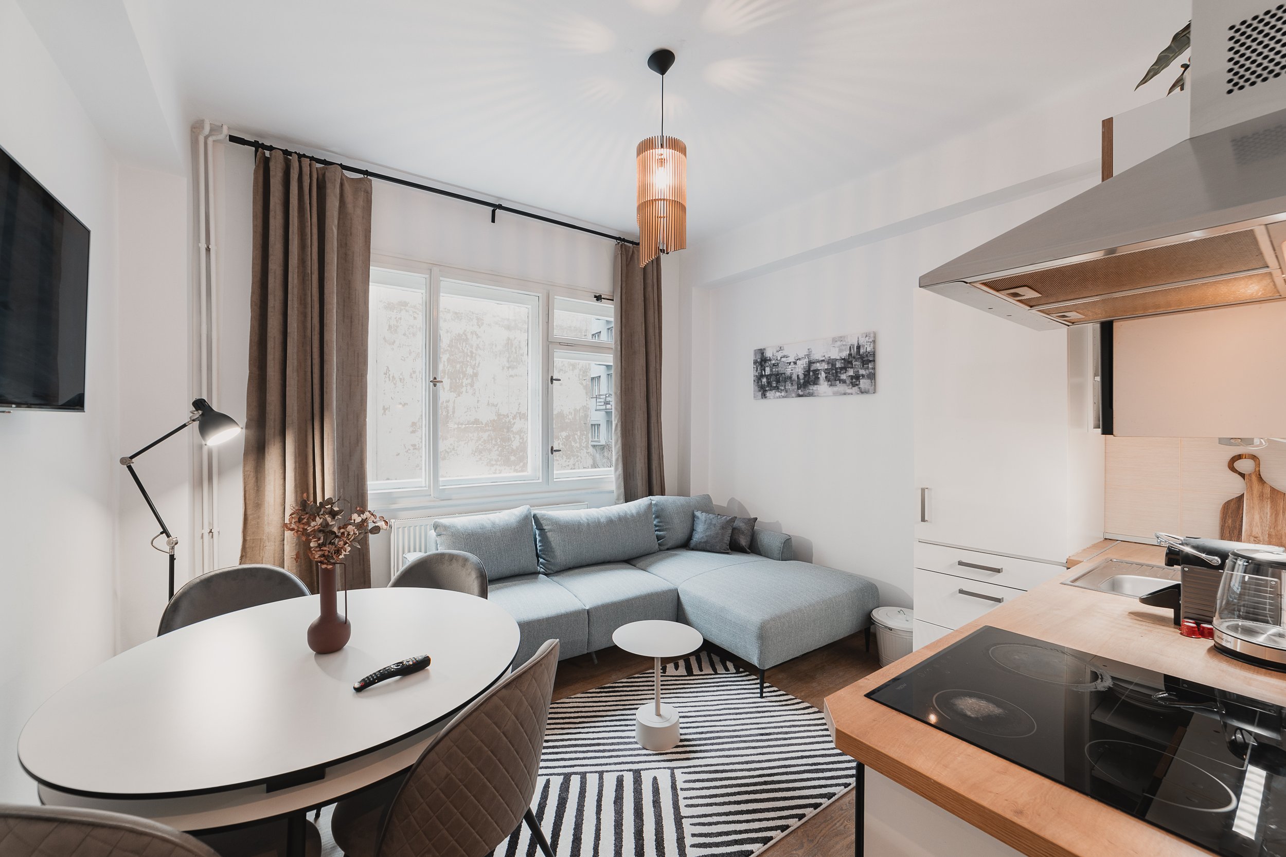 Living room with large windows, beige curtains, gray sofa, black and white artwork, white kitchen cabinets, black stovetop, round dining table with four beige quilted chairs, striped black and white rug, and a pendant light fixture.