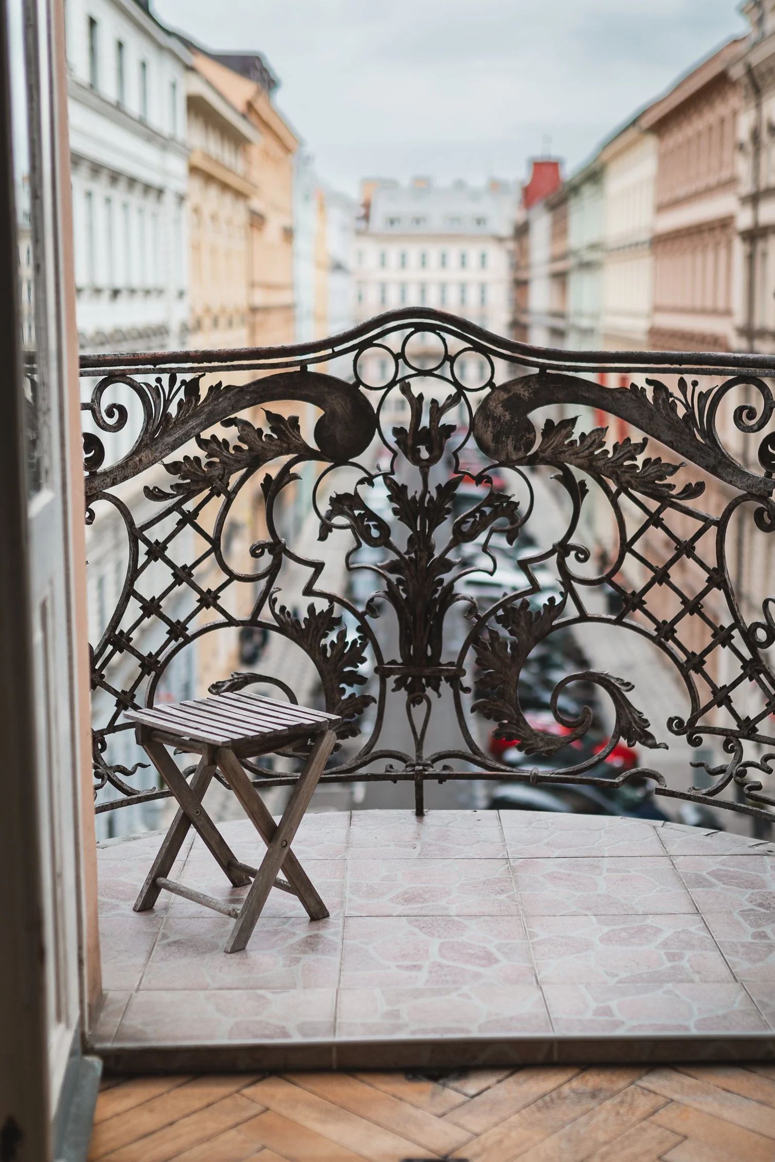 View from a balcony showing an ornate iron railing, a small wooden table and chair, with buildings across the street and a cloudy sky in the background.