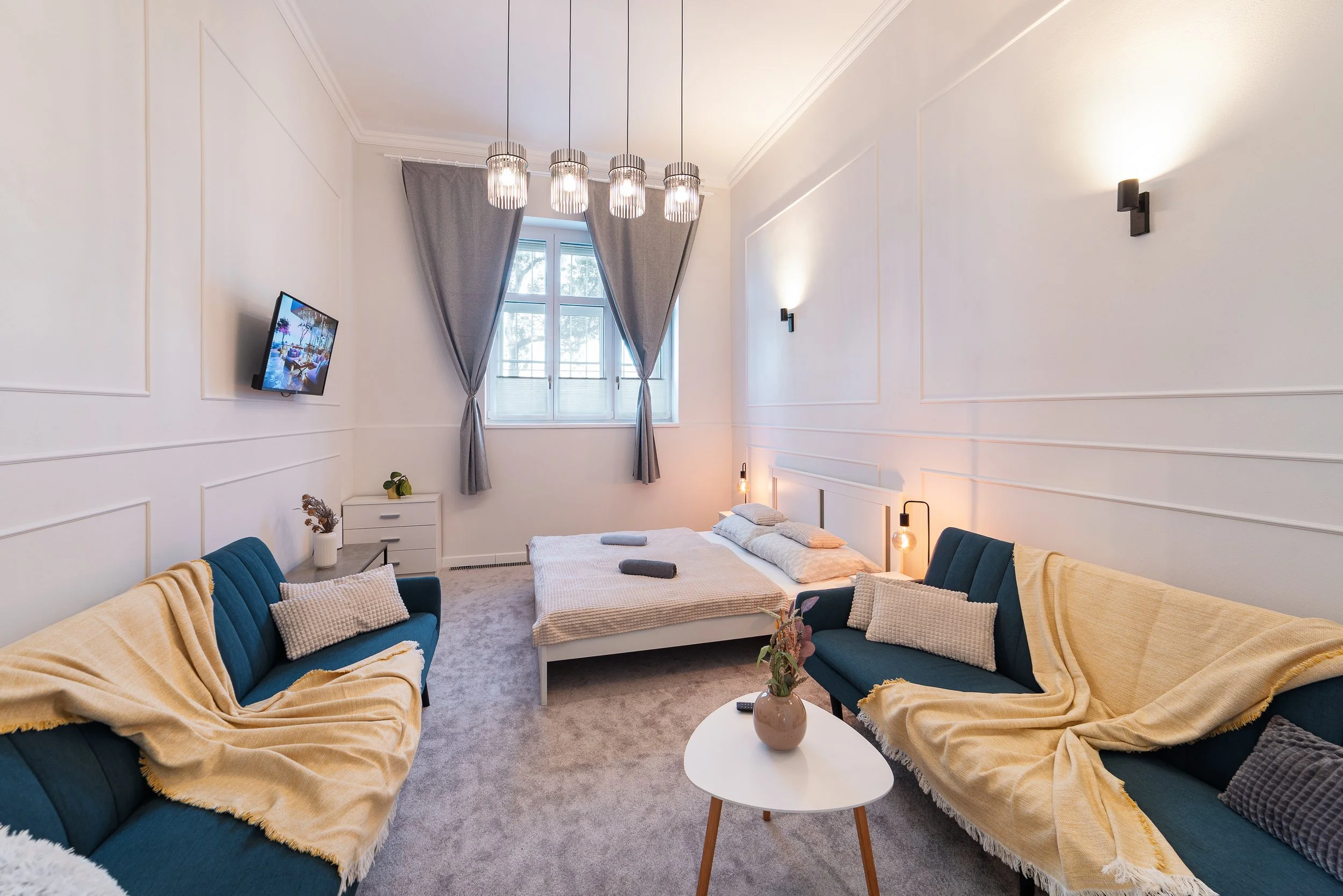 A modern bedroom with white paneled walls, a large window with gray curtains, a central bed, and a seating area around it with blue sofas and yellow throws. There is a television on the wall, a small white side table, and several light fixtures inclu