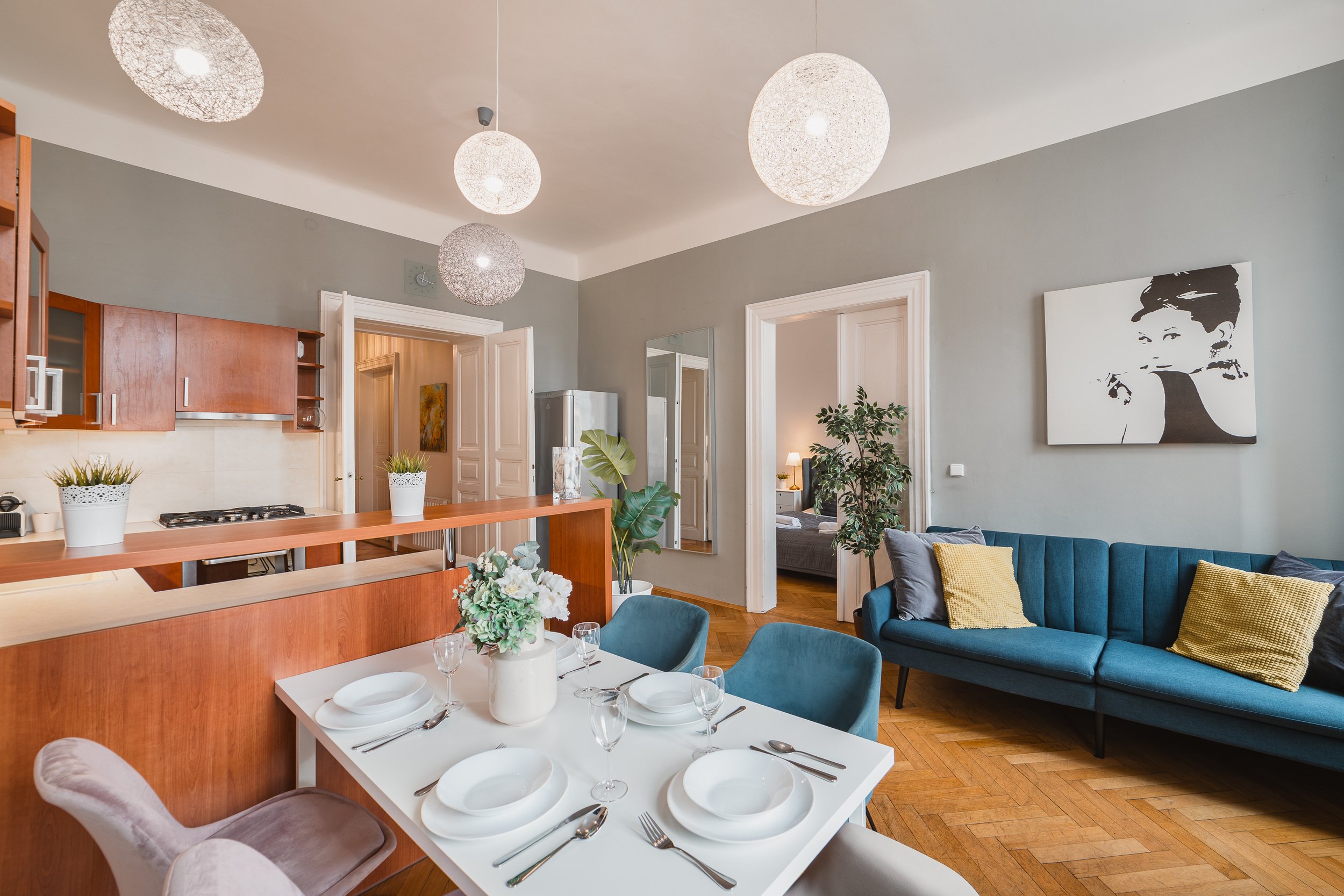 Bright living and dining area with a blue sofa, dining table set for six, decorative plants, wall art, and modern pendant lights.