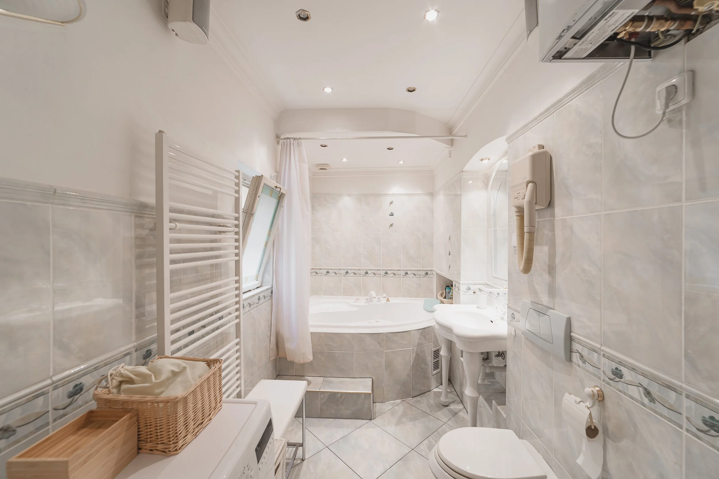 A small, narrow bathroom with white tiled walls and ceiling, featuring a bathtub with a shower curtain, a white toilet, a white sink, a wicker basket on a white surface, a white towel heater, an electronic device holder, and a small window with a whi