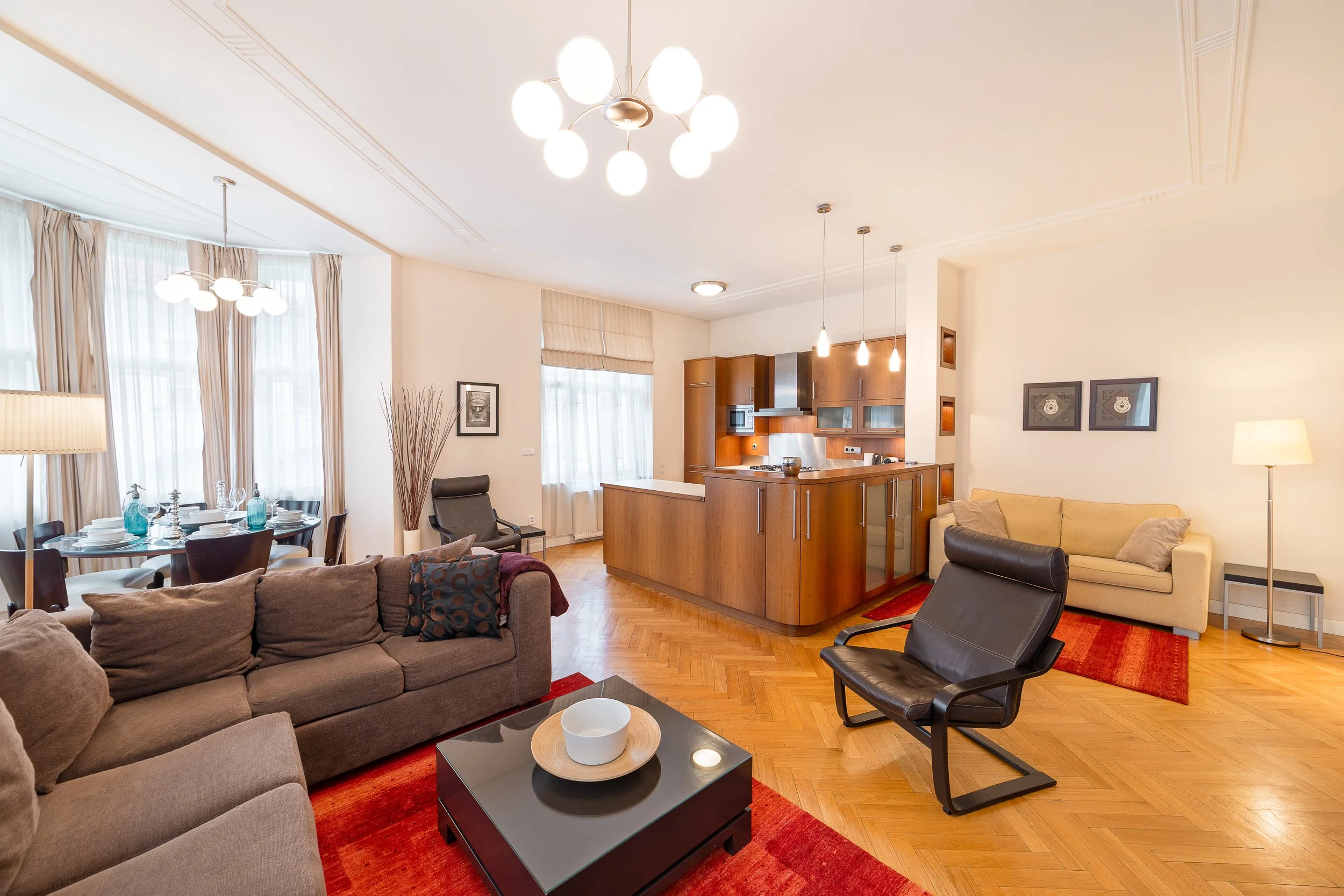 Living room with a beige sofa, black lounge chair, coffee table with decorative bowl, red area rug, multiple pendant and ceiling lights, wooden kitchen cabinets, dining table with chairs, large windows with sheer curtains, and wall art.