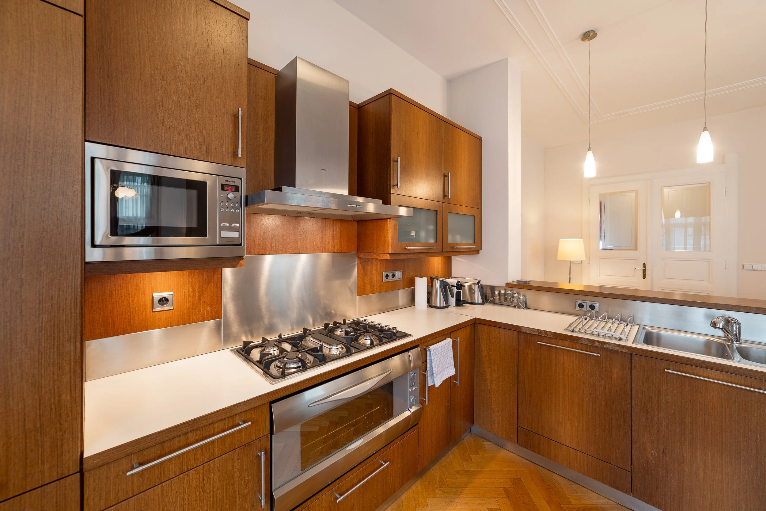 Modern kitchen with wooden cabinets, stainless steel appliances including microwave, oven, and range hood, and a countertop with various small appliances and utensils. There are pendant lights and a door with windows in the background.