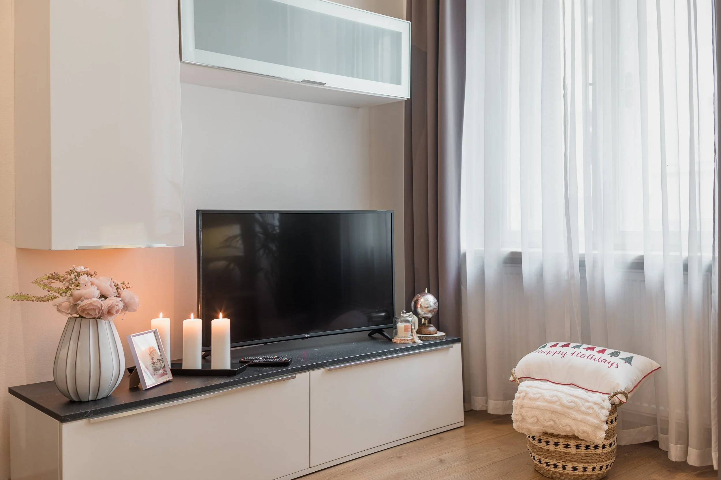 Living room entertainment area with a flat-screen TV on a white cabinet, decorated with lit candles, a framed photo, a flower vase with pink flowers, and a small globe. A round wicker basket with folded towels and a holiday pillow with 'happy holiday