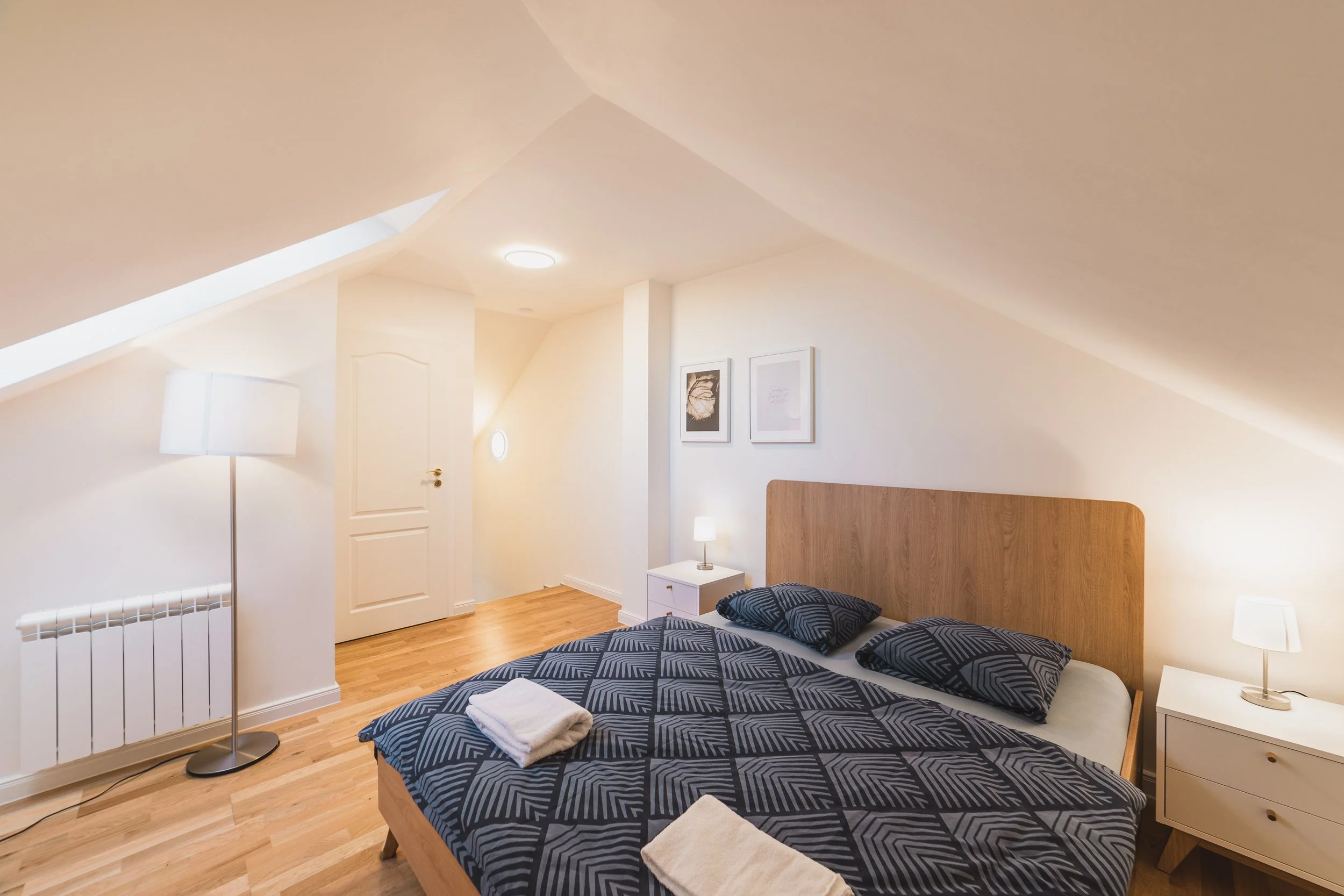 A bedroom with a bed featuring dark blue geometric patterned bedding, a white nightstand holding a small lamp, and wall art. The room has sloped ceilings, hardwood floors, and a wooden headboard. There is a floor lamp and framed pictures on the wall.