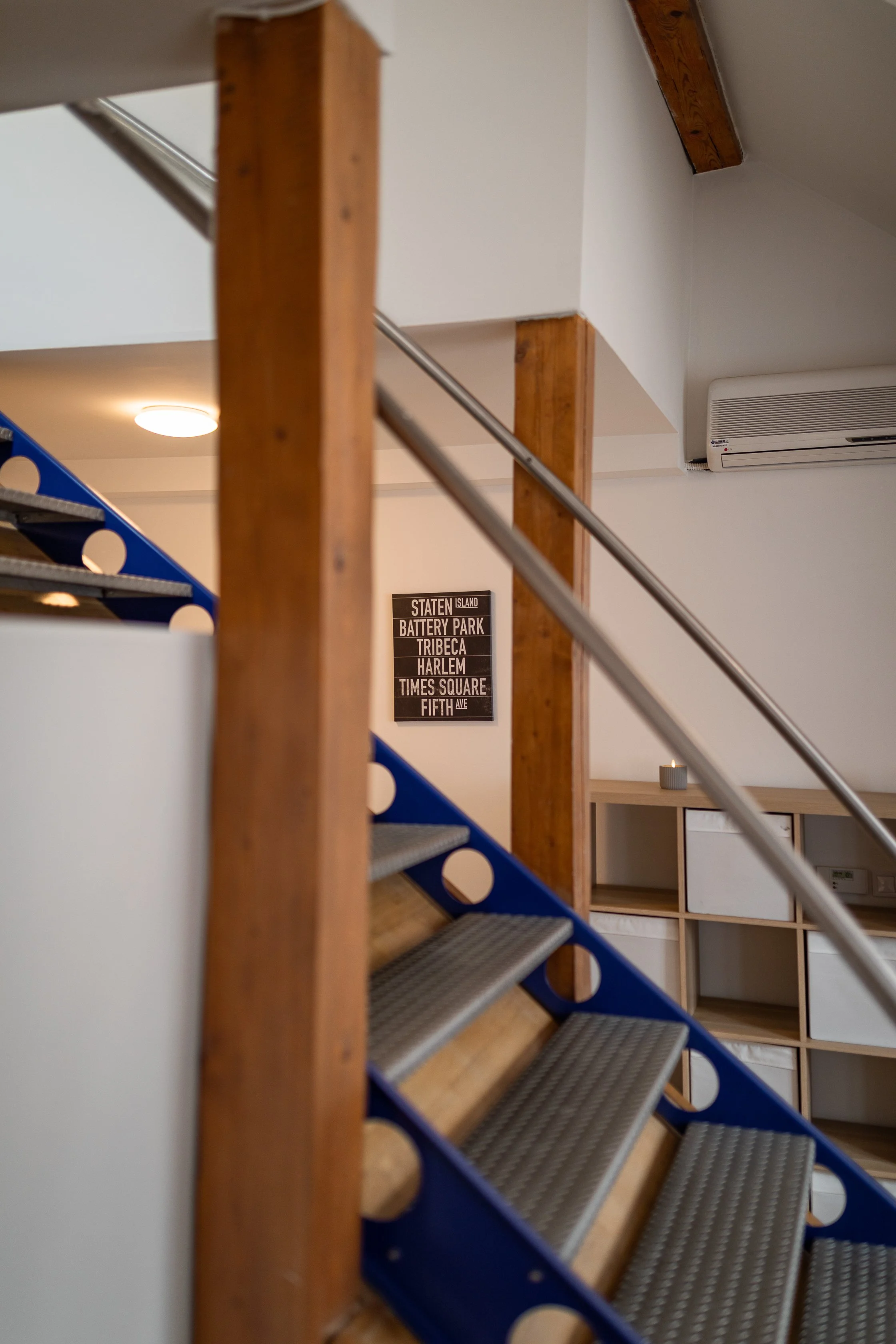 Indoor staircase with wooden railing, metal steps, and a wall-mounted air conditioner in the background. A poster listing New York City locations is visible on the wall.