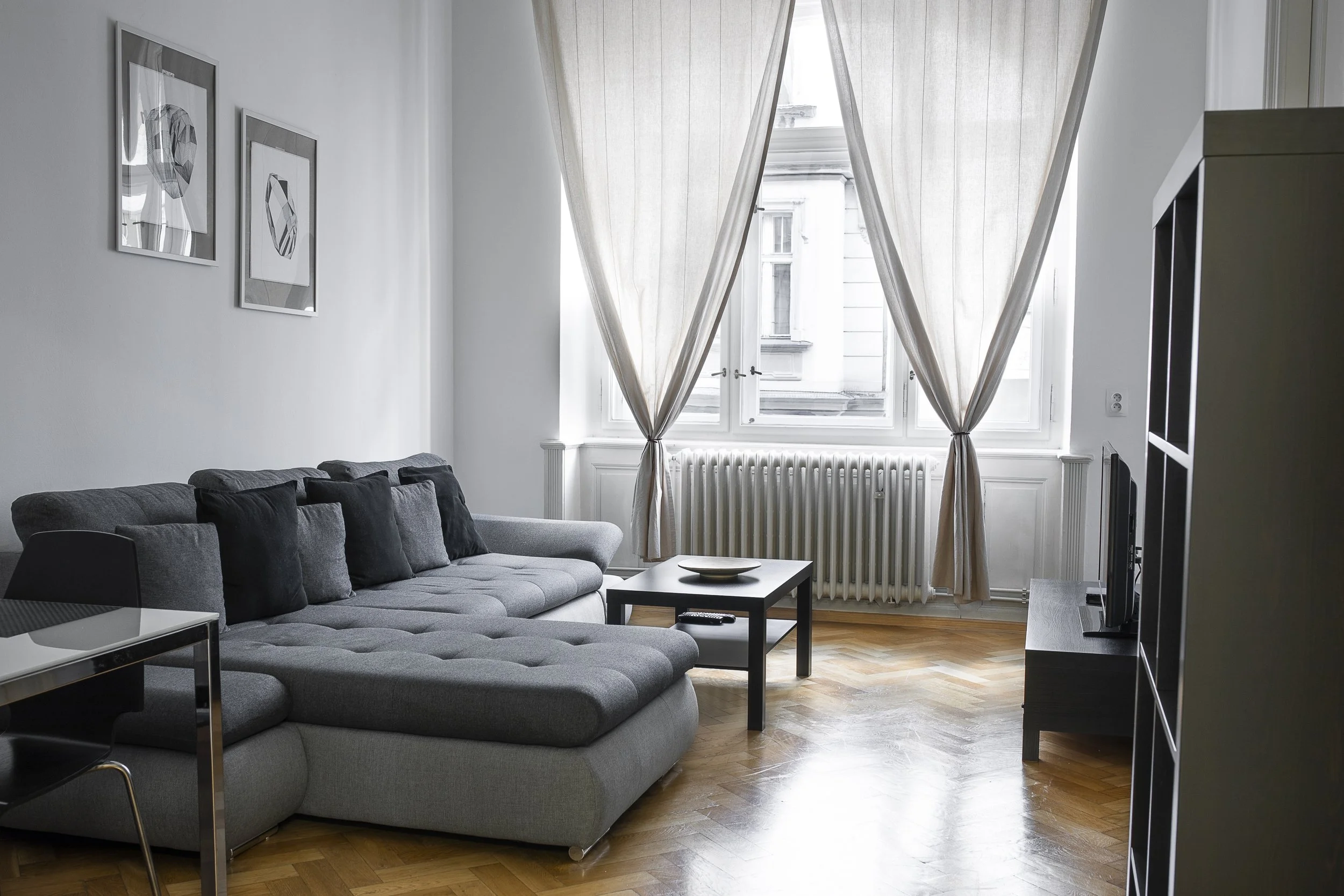 Living room with a large gray sectional sofa, black pillows, glass side table, TV on a black stand, large window with sheer beige curtains, artwork on the white wall, and hardwood flooring.