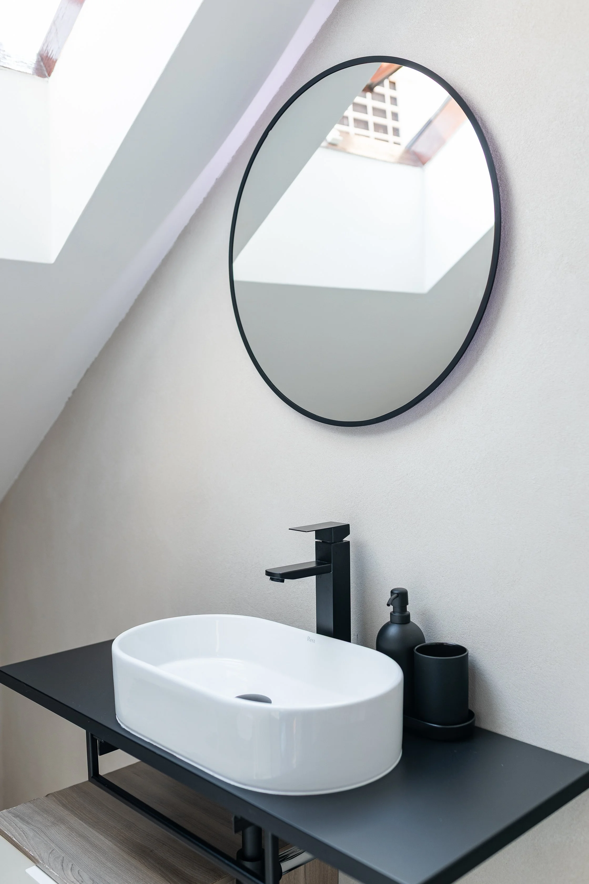 Modern bathroom with black countertop, white vessel sink, black faucet, black soap dispenser, and a round mirror on a white wall with skylights.