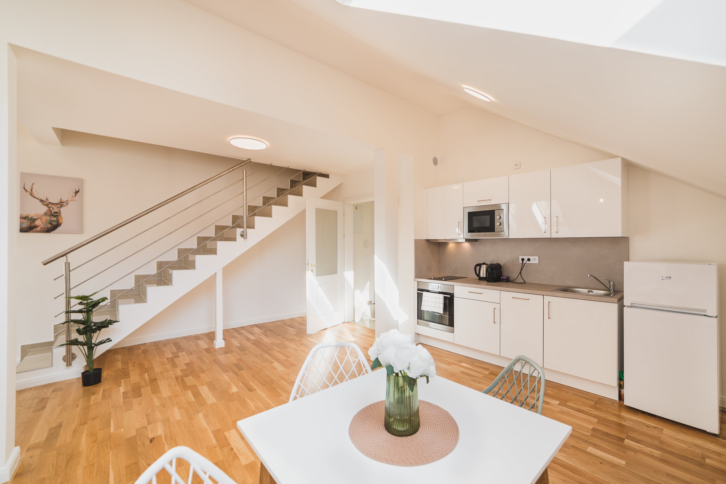 Interior of a modern apartment with a white kitchen, wooden flooring, a dining table with white chairs, a vase with white flowers, a staircase with a metal railing, a framed deer portrait on the wall, and a door leading to another room.