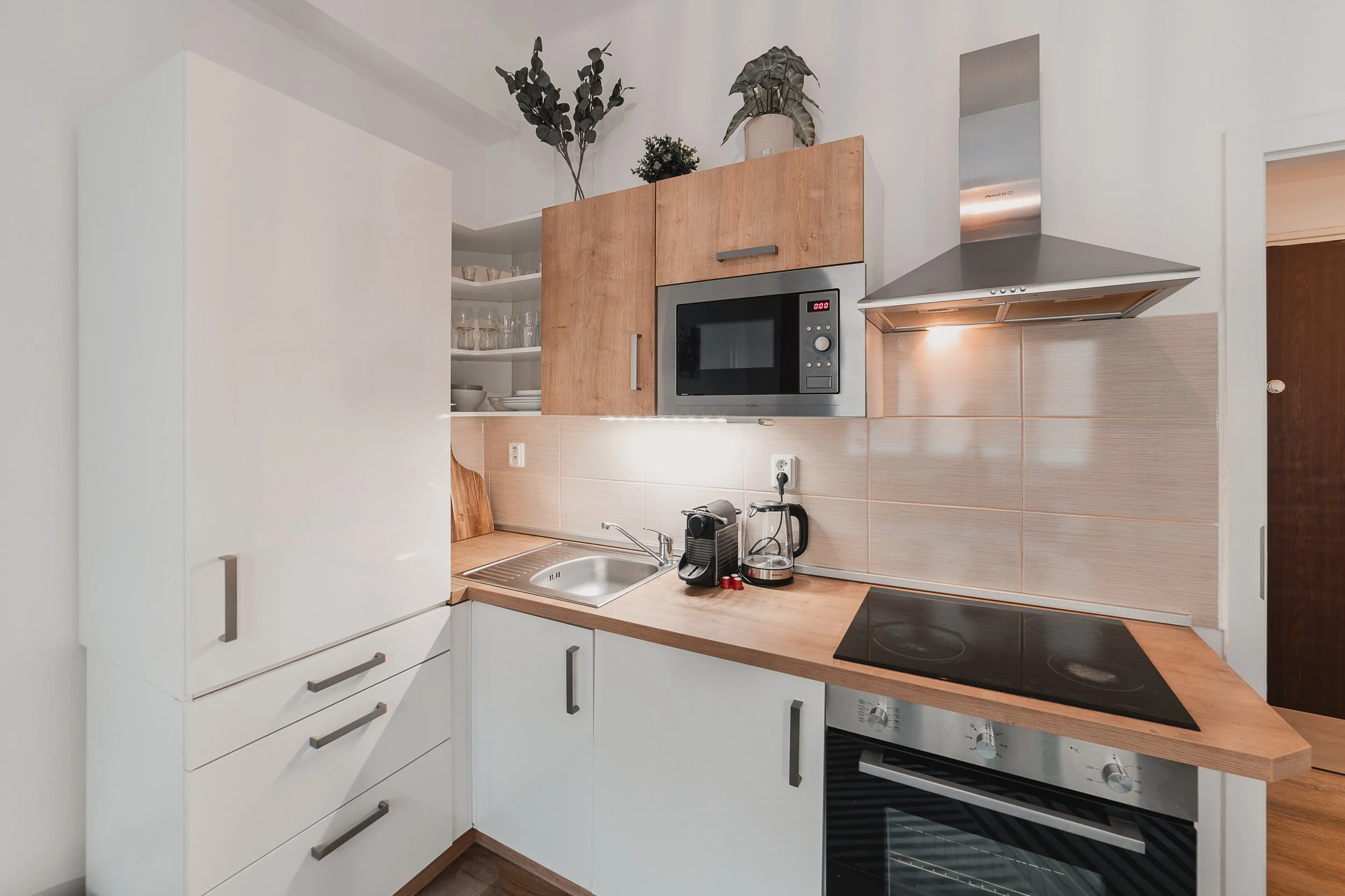 Modern kitchen with white cabinets, wooden countertop, and built-in microwave. Includes a sink, coffee machine, and stovetop with oven. Decor includes potted plants on top of upper cabinets.