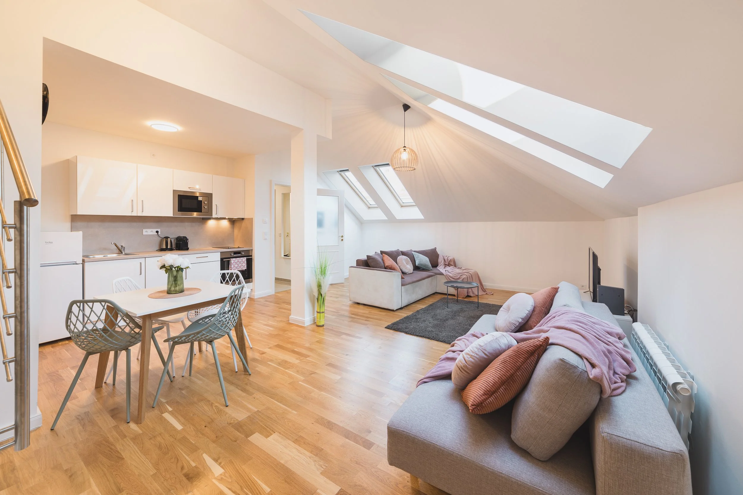 Open-concept living space with kitchen, dining, and sitting area. Light wood floors, white cabinets, and skylights illuminate the room. Furnished with sofas, a dining table, and decorated with pillows and plants.