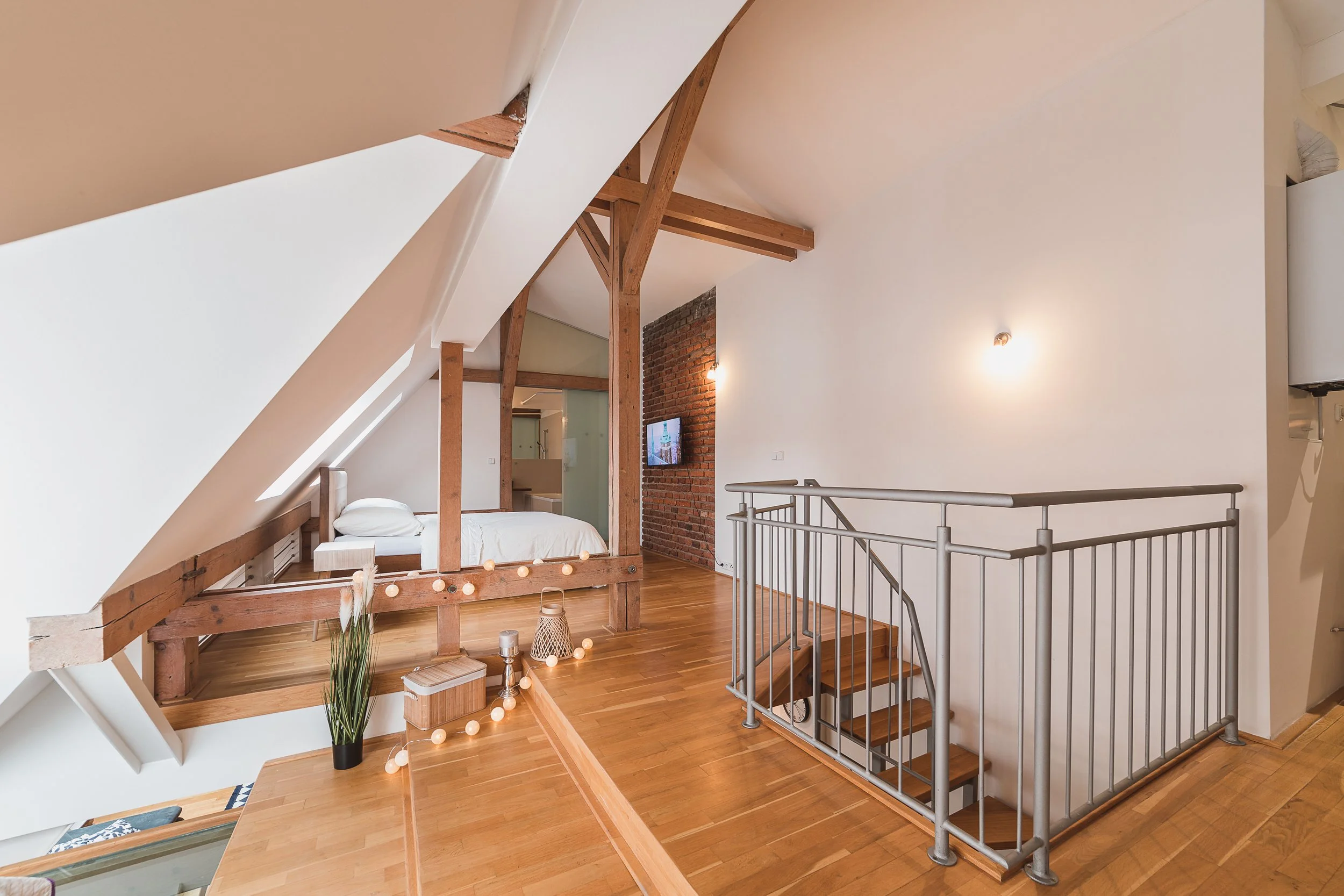 Interior of an attic space with a sloped ceiling, wooden and brick accents, a bed with white bedding, decorative lights, a potted plant, and a metal railing around a staircase.