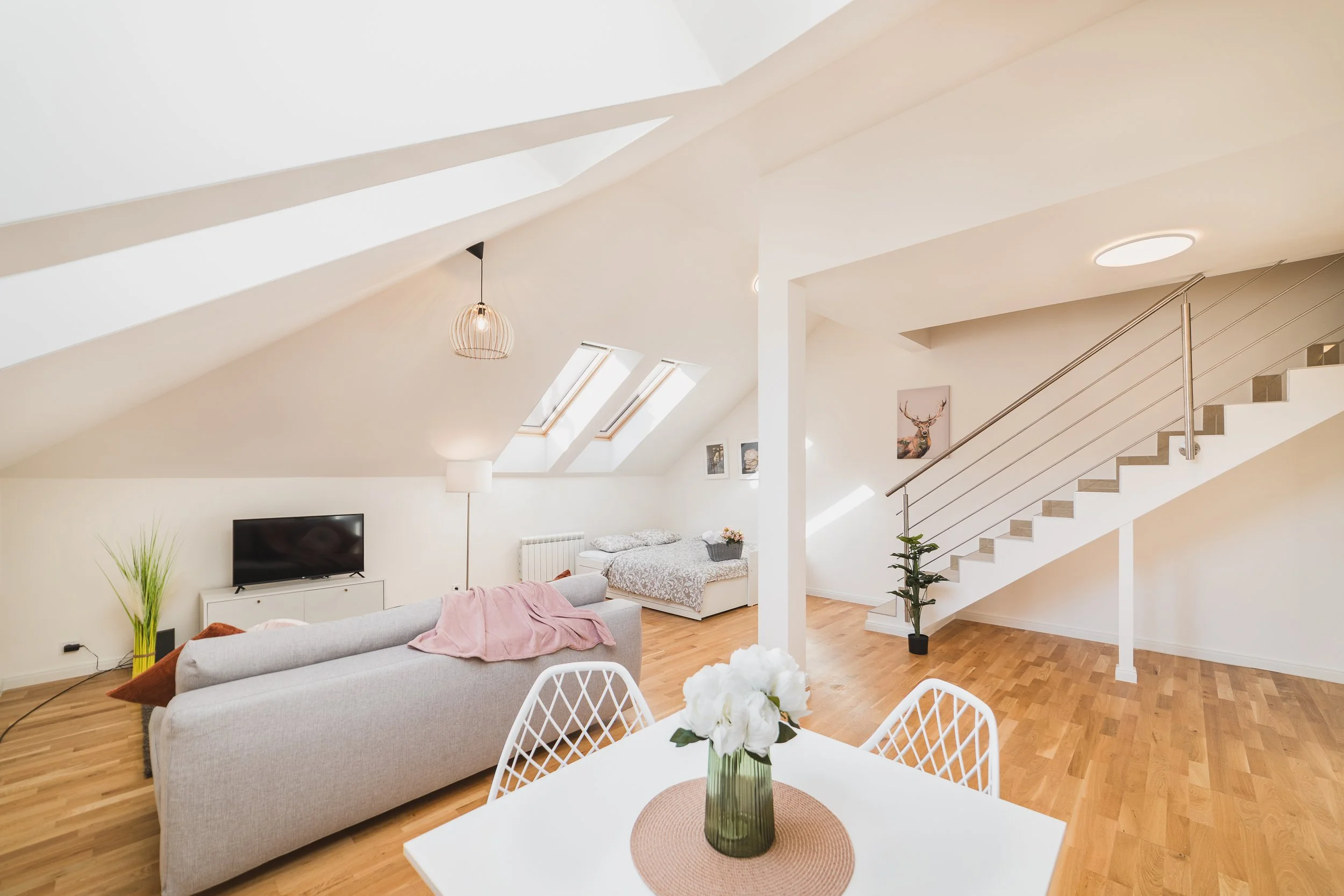 A spacious living room with white walls and ceiling, hardwood floors, skylights, a modern hanging light fixture, a gray sofa with pink blanket, a white TV stand with a television, framed artwork on the wall, a staircase with metal railing, and potted