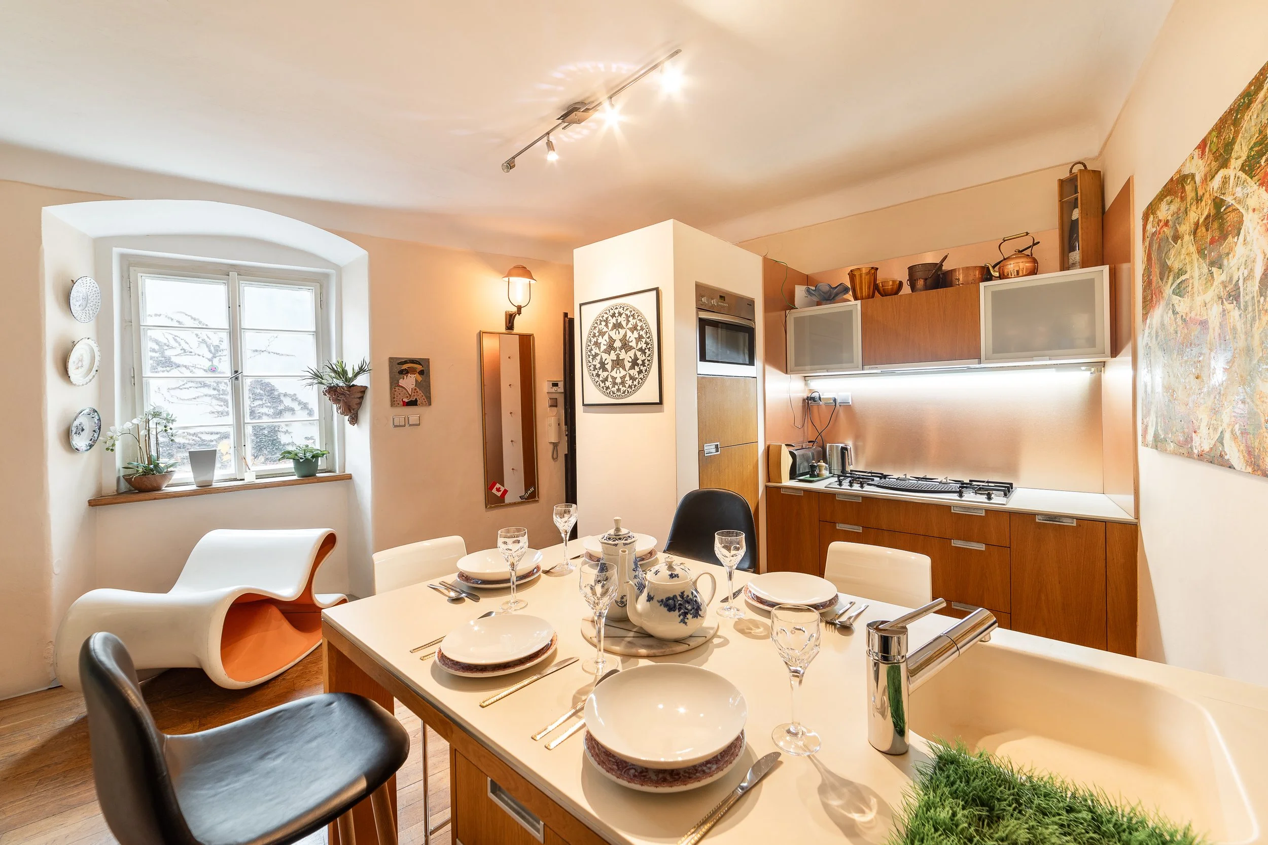 A dining area with a table set with plates, glasses, and teapots, surrounded by modern chairs. The background shows a kitchen with wooden cabinets, a window with plants, and artwork on the walls.