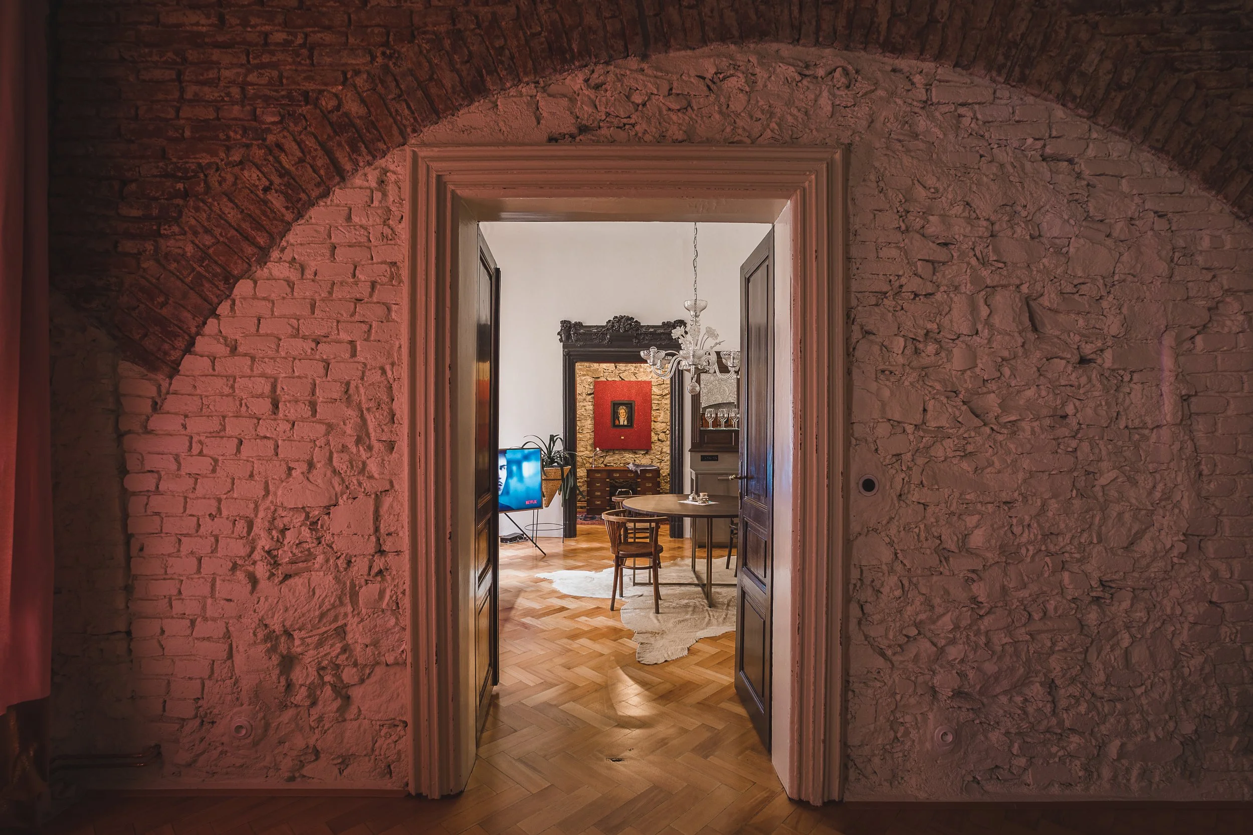 View through a doorway into a living room with a chandelier, a painting on the wall, and a hat on a chair on a rug with wooden floor.