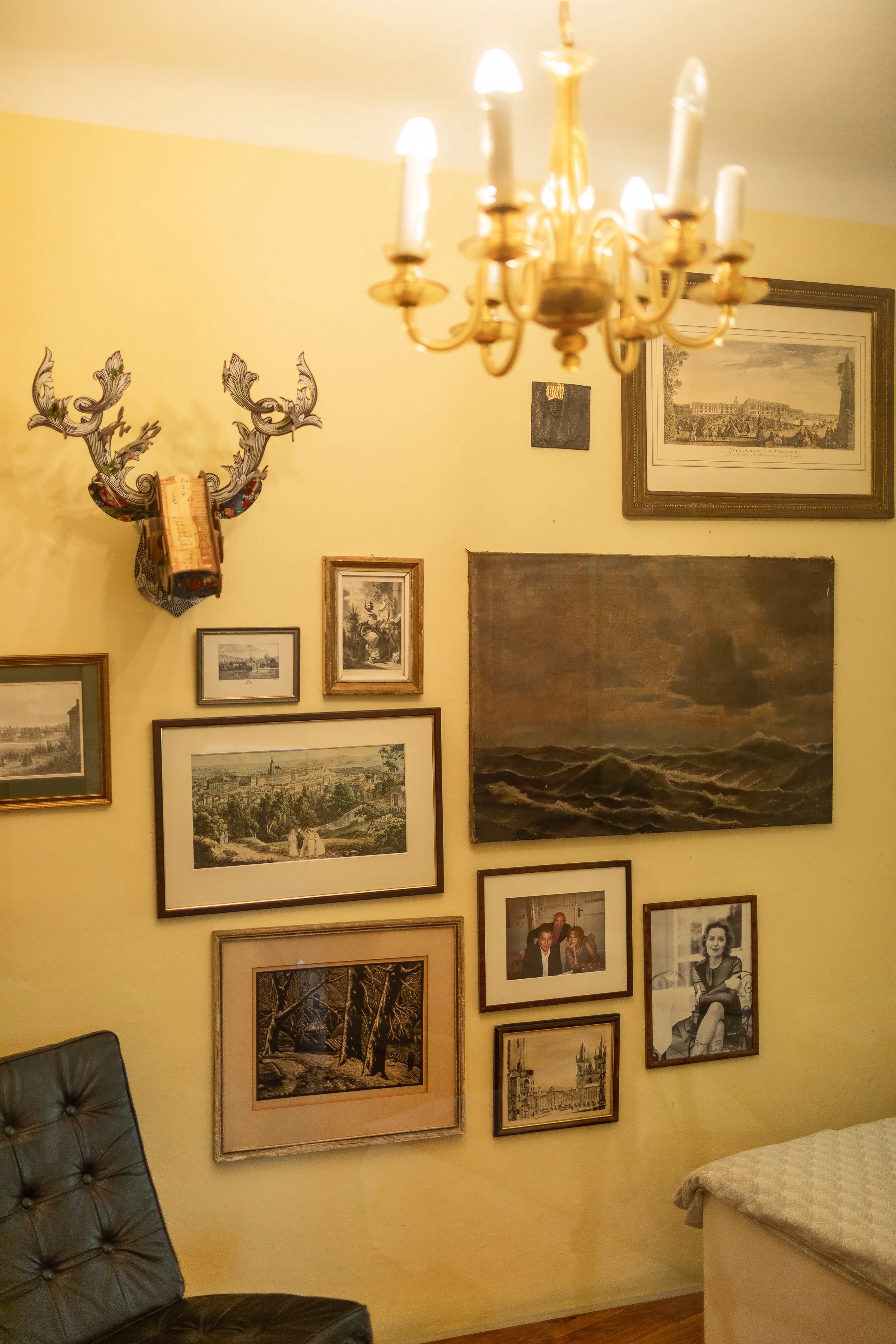 A living room corner with a yellow wall decorated with various framed pictures and artwork, including landscape paintings and photographs. A brass chandelier with candle-style bulbs hangs from the ceiling. A decorative deer head with colorful accents