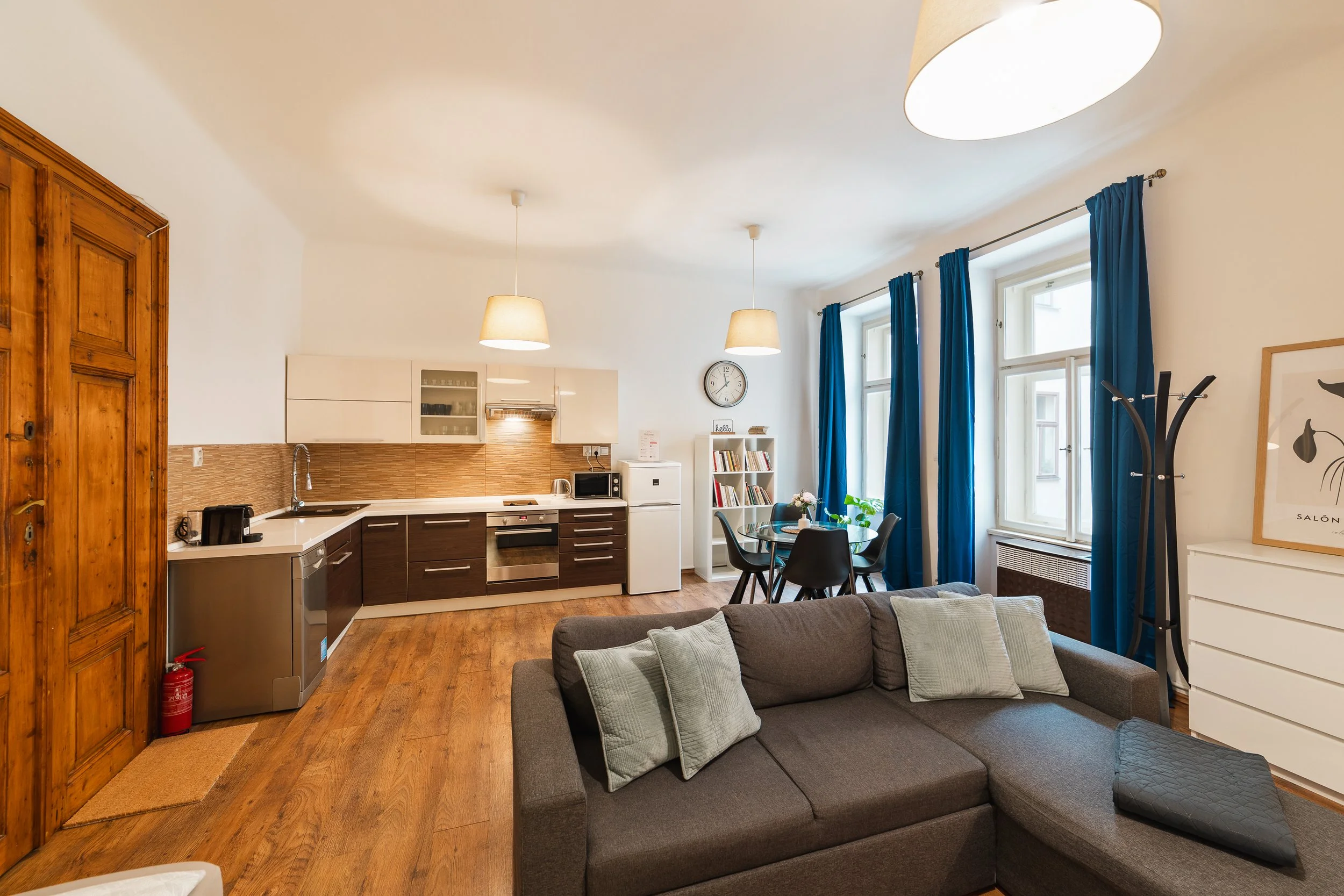 Open-concept living room and kitchen with hardwood floors, a gray sofa with pillows, a round dining table with chairs, large windows with blue curtains, and white walls decorated with a clock and a framed artwork.