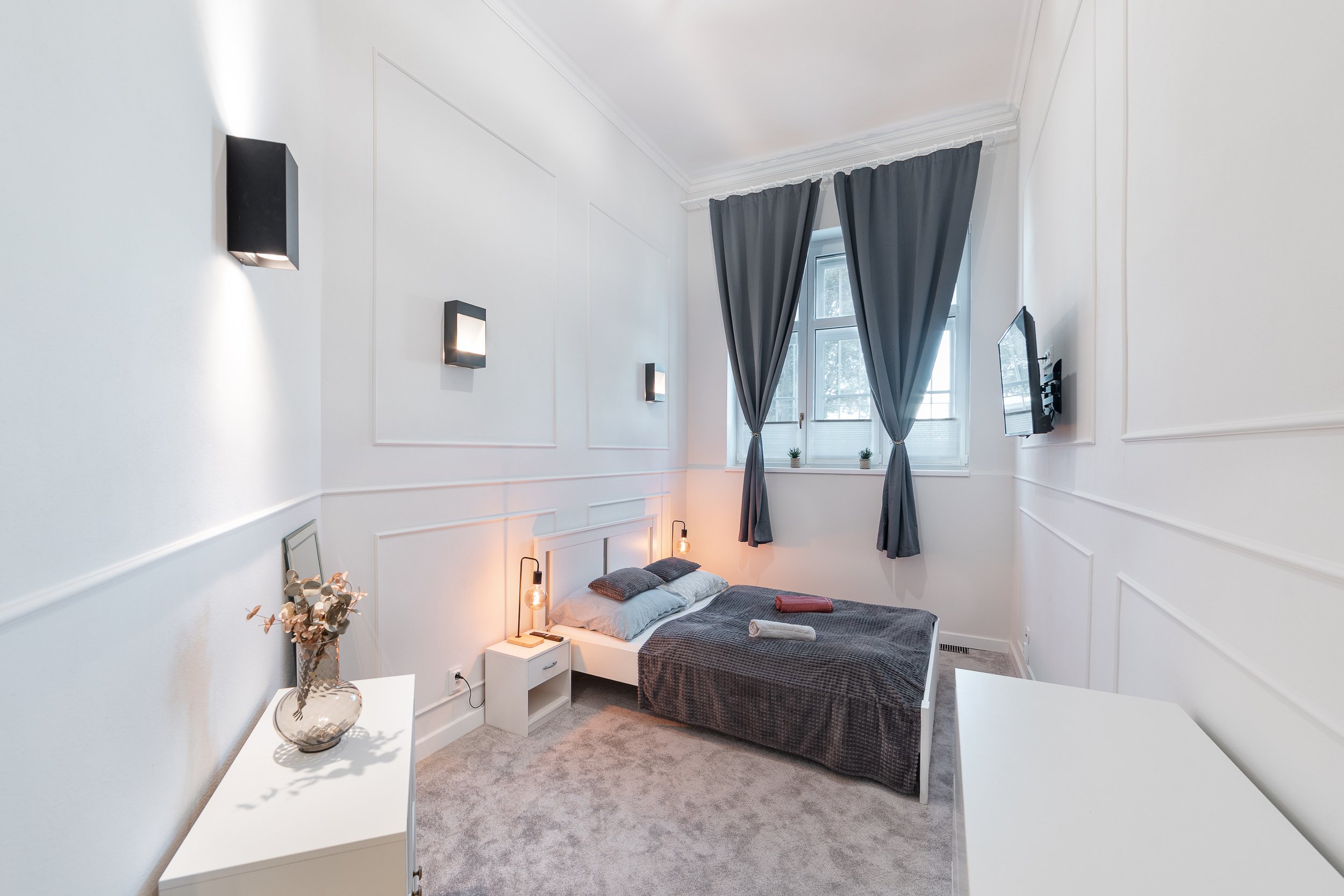 A bedroom with white walls, a bed with dark gray bedding and pillows, two pendant lights hanging over the bed, a bedside table with a book and a lamp, a window with gray curtains, and wall-mounted black lights.