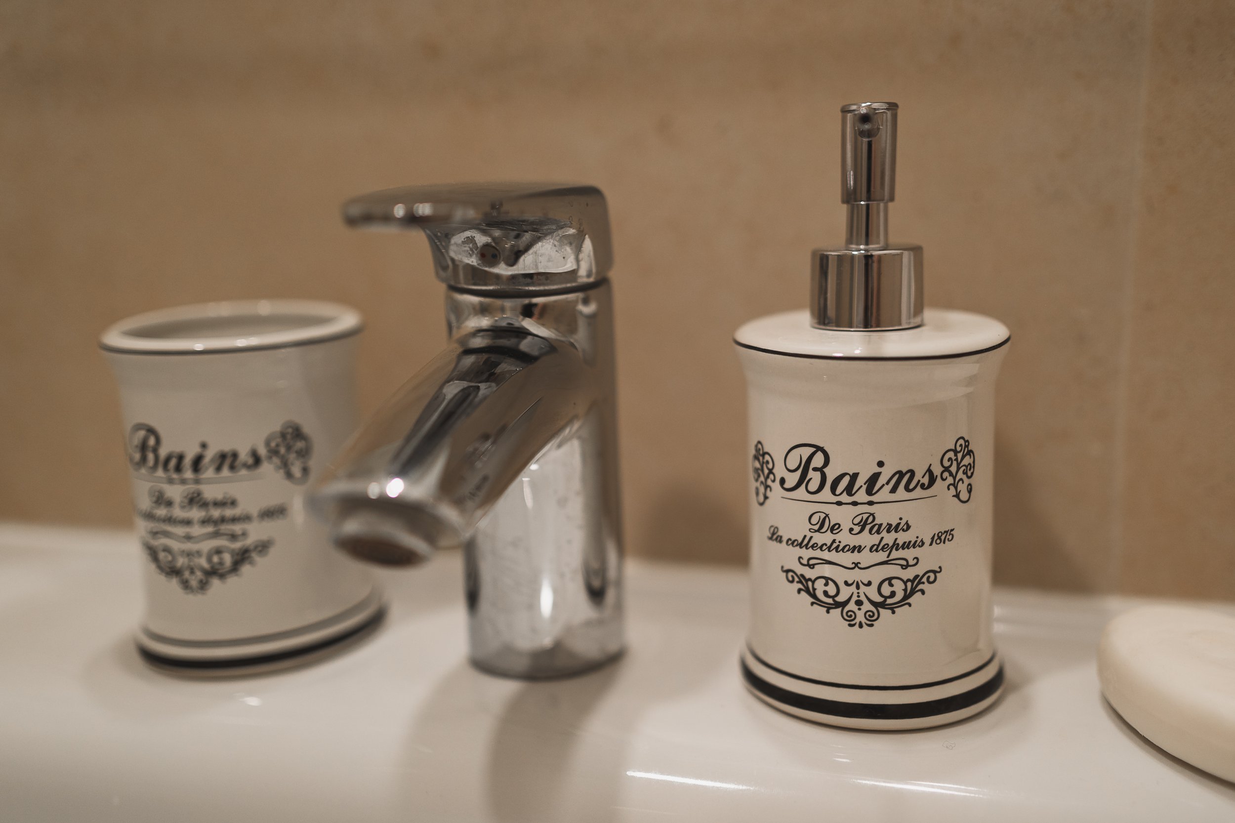 A bathroom sink with a chrome faucet, a cream-colored soap dispenser, and a matching cream-colored cup, all with black decorative writing and designs.