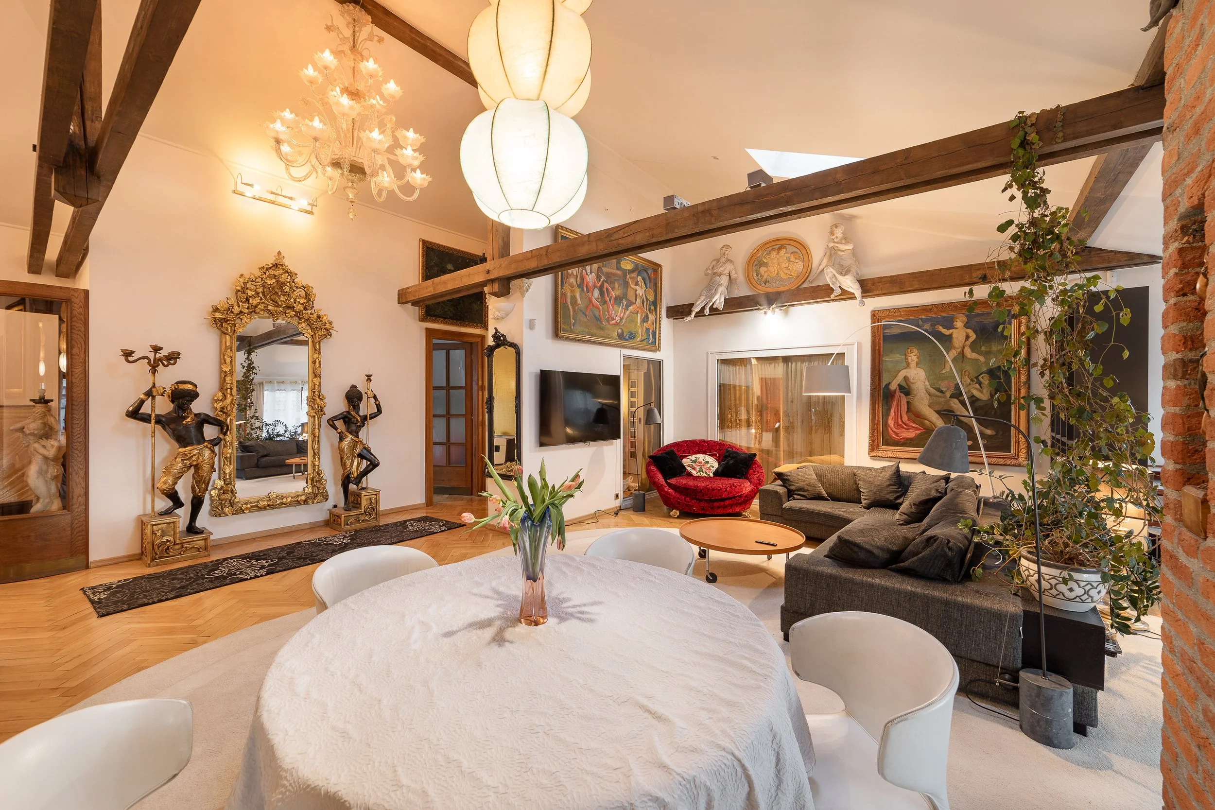 A spacious living room with high ceilings, exposed wooden beams, and eclectic decor. Features include a chandelier, large paintings, sculptures, a flat-screen TV, and a mix of vintage and modern furniture. There's a round dining table with a vase of 