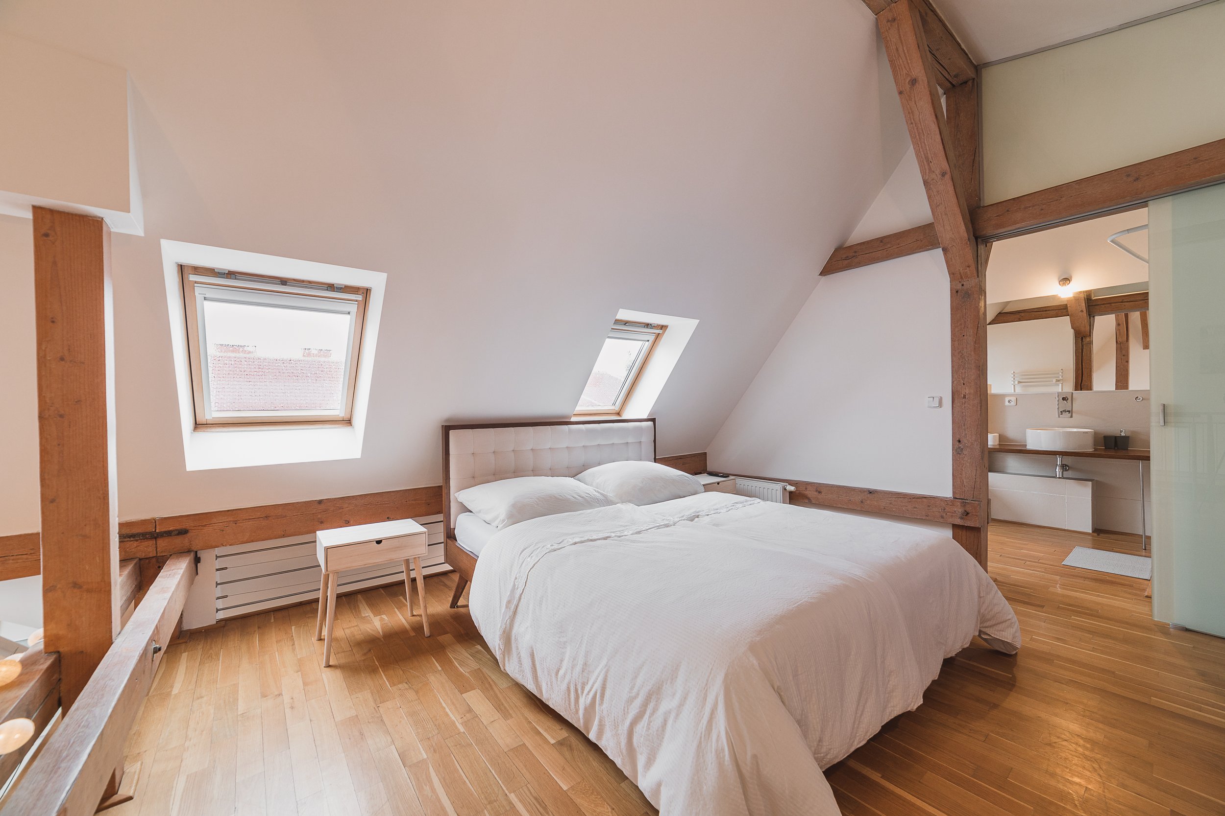 A cozy attic bedroom with a sloped ceiling, two skylight windows, a white bed with a wooden frame, a small white nightstand, and exposed wooden beams.