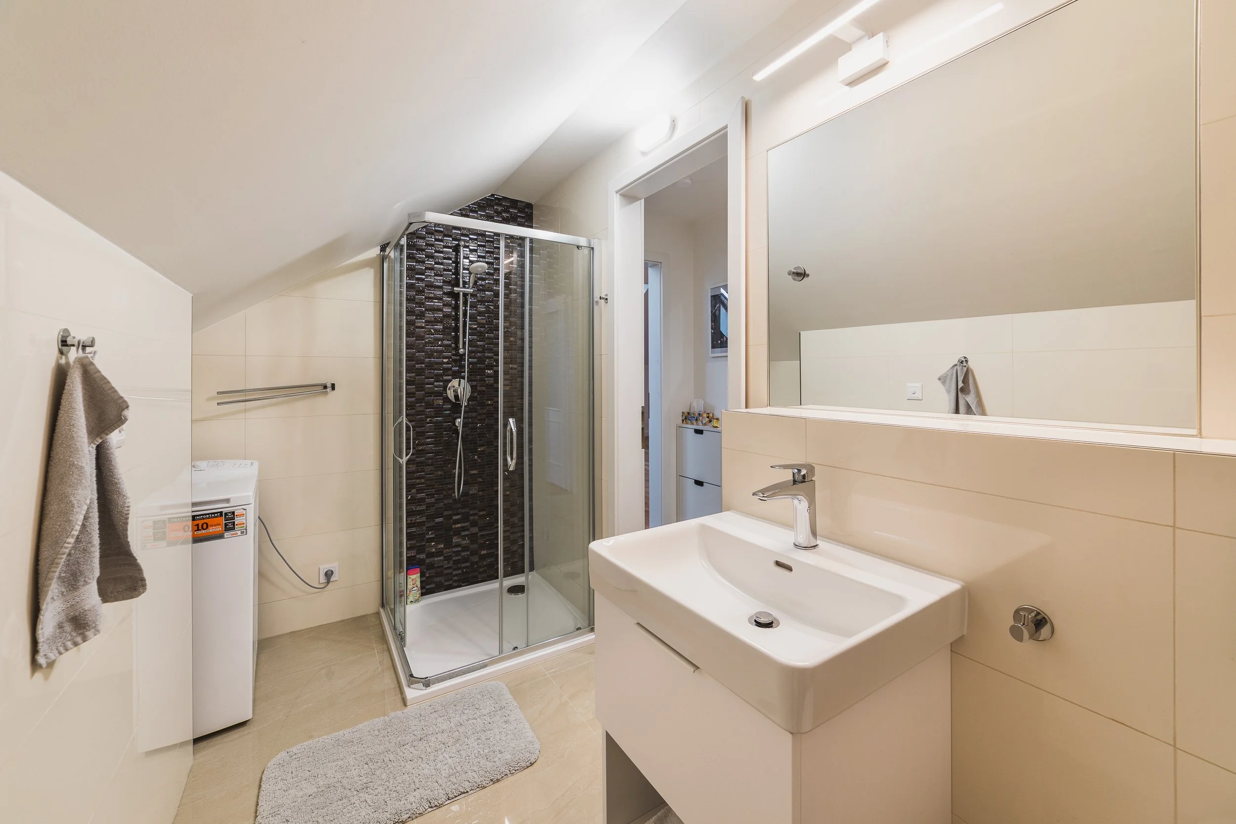 A modern bathroom with a small shower enclosure with dark mosaic tiles, a white laundry machine, a white sink with a mirror, beige tiled walls, and a towel hanging on a wall hook.