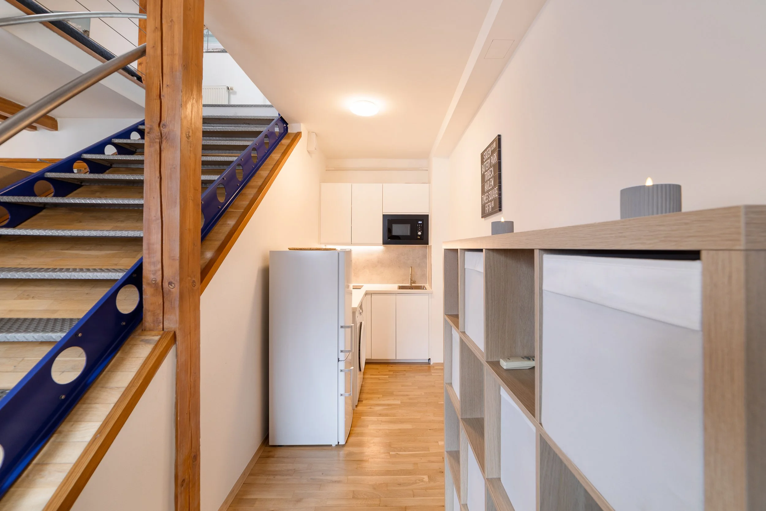 Interior view of a small apartment kitchen with a white refrigerator, microwave, and wooden shelving unit. A staircase with wooden and blue metal railings leads to an upper level. The space features light wood flooring, white walls, and simple, moder