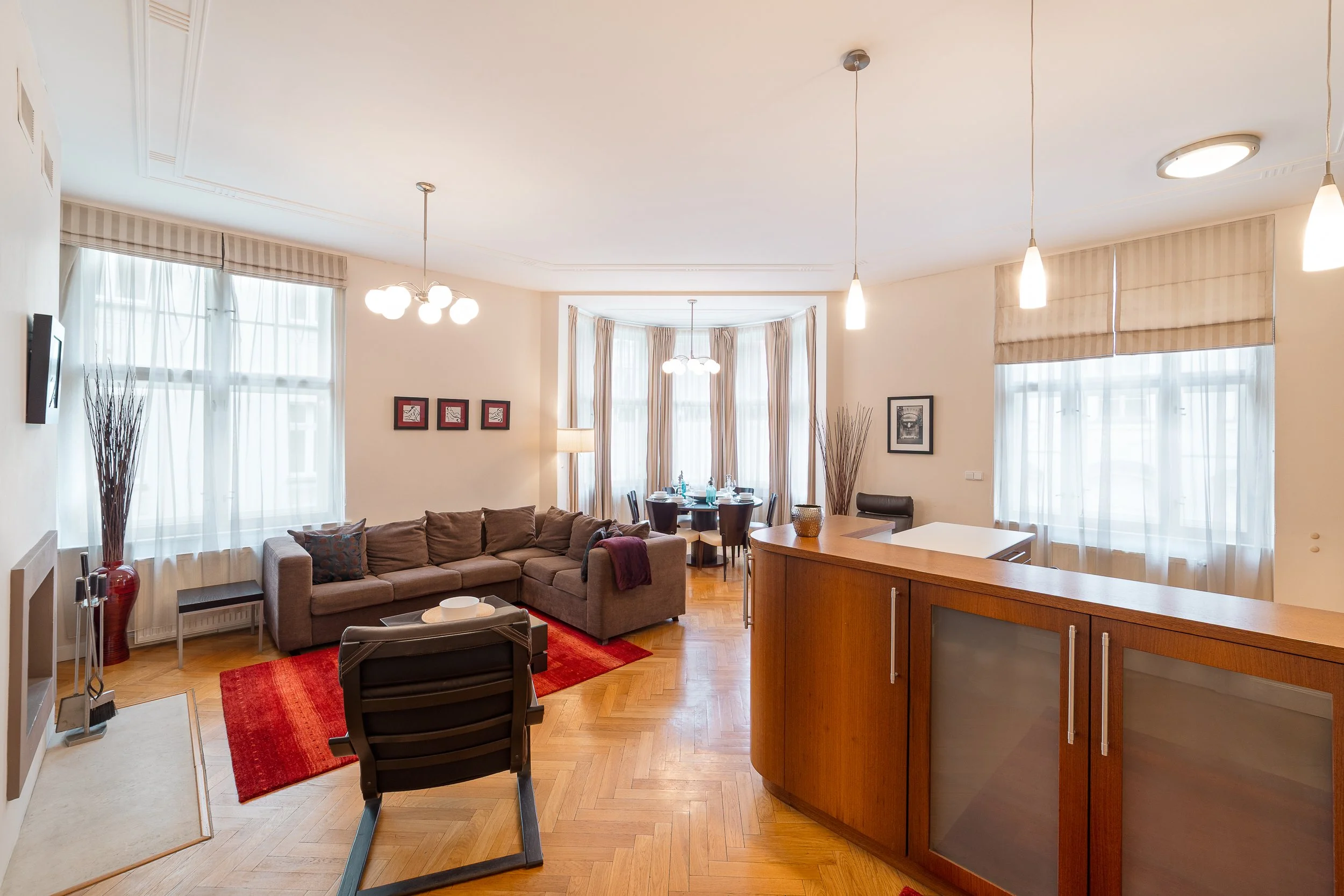 A spacious living room with a beige sectional sofa, dark wooden chairs, a small side table, and a red area rug. Large windows with sheer curtains allow natural light, and the ceiling has modern pendant and chandelier lighting fixtures. The room also 