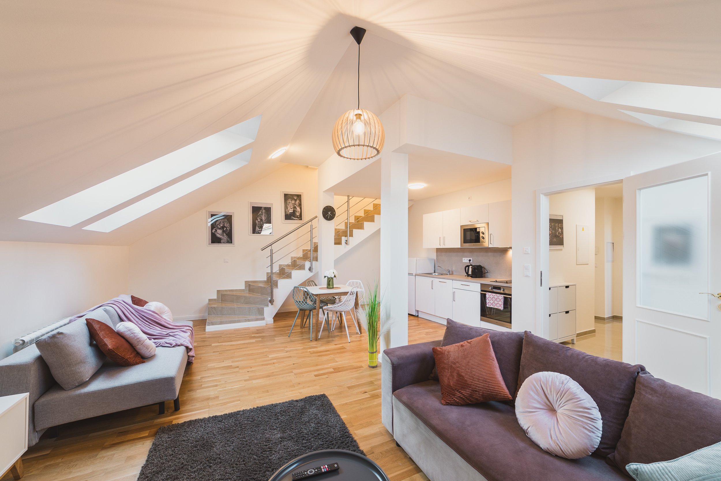 Open-concept living space with a vaulted ceiling, skylights, and modern furniture including two gray sofas with pillows, a small dining table with chairs, and a staircase leading to an upper level. The room has wooden flooring, wall art, and a kitche