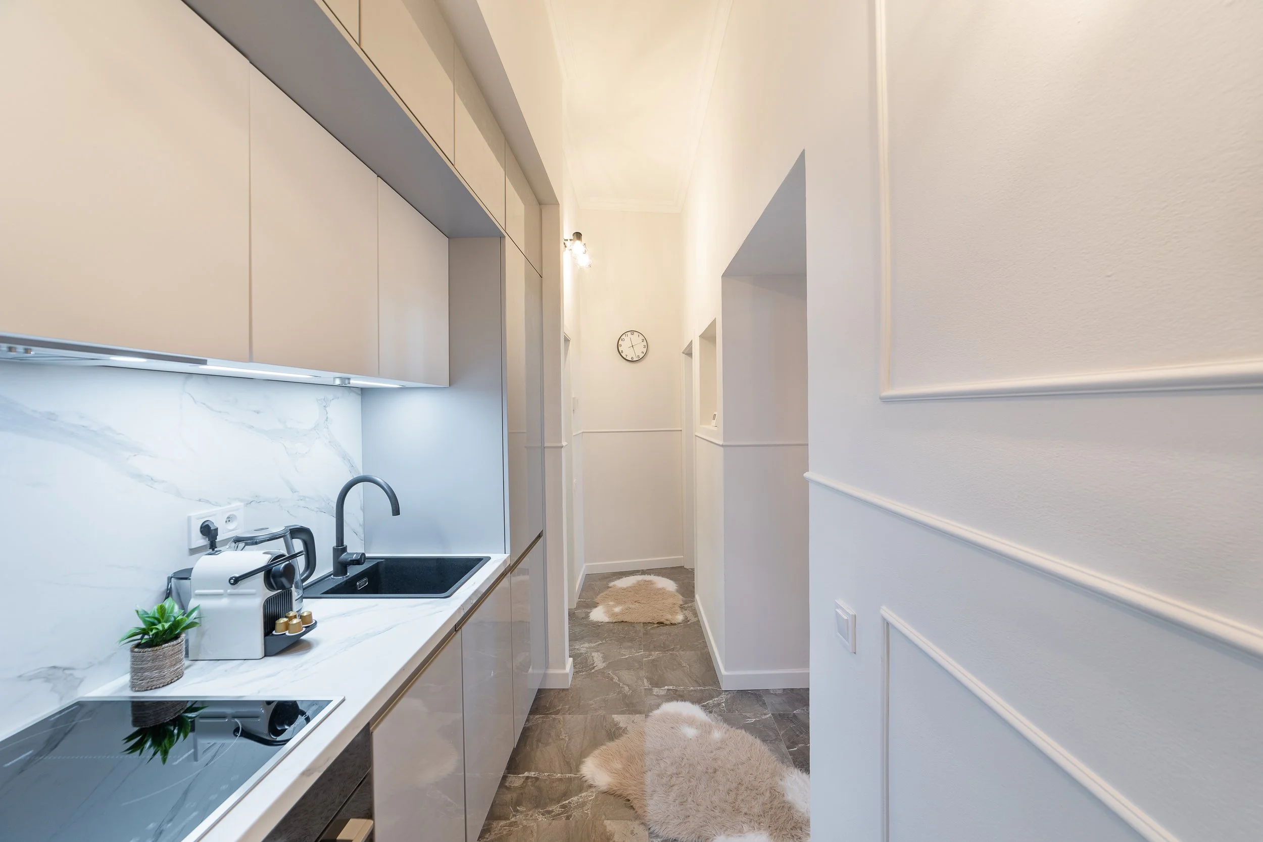 A modern kitchen with white cabinets, a marble countertop, and a small sink. There is a potted plant on the counter, and a wall clock on the far wall. The hallway next to the kitchen has white walls with decorative molding and a beige furry rug on th