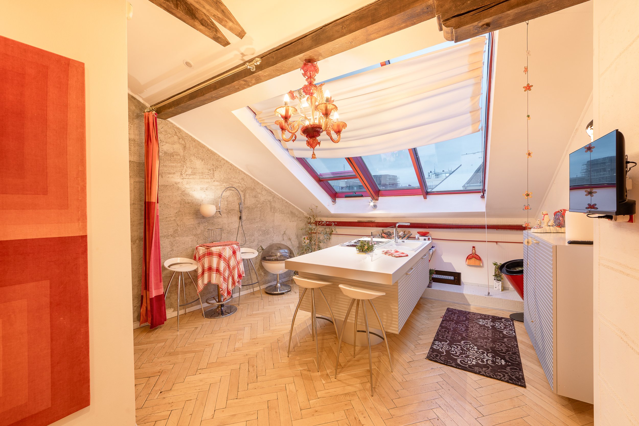 An attic-style kitchen and dining area with a sloped ceiling, exposed wooden beams, a chandelier, a large skylight window, and a sectional white kitchen island with barstools. There are plants and kitchenware on and around the island, and colorful cu