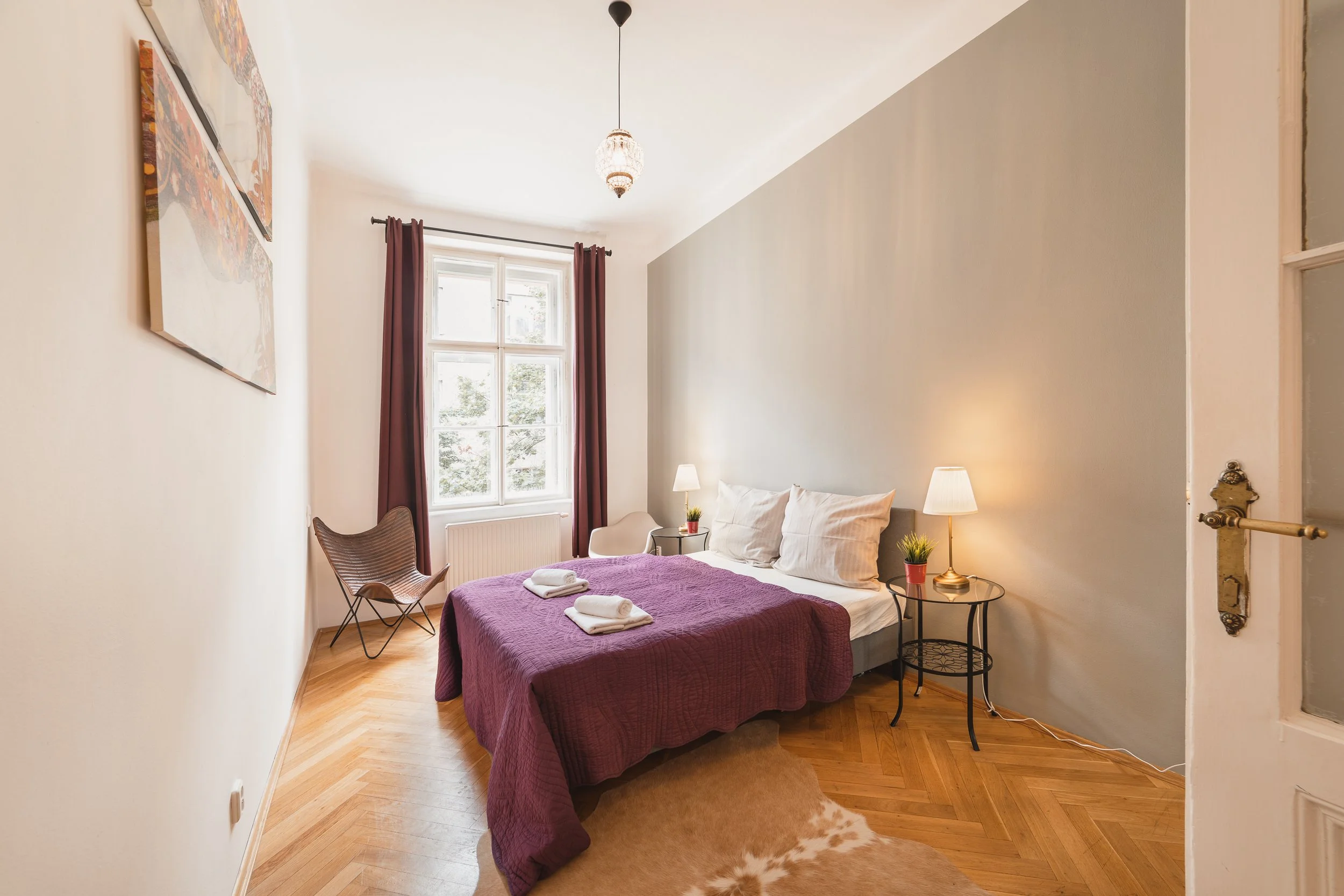A cozy bedroom with a double bed covered in a purple quilt, white pillows, and two neatly folded towels. The room has wooden flooring, a window with maroon curtains, and a small black side table with a lamp and plant. There are two chairs, one wicker