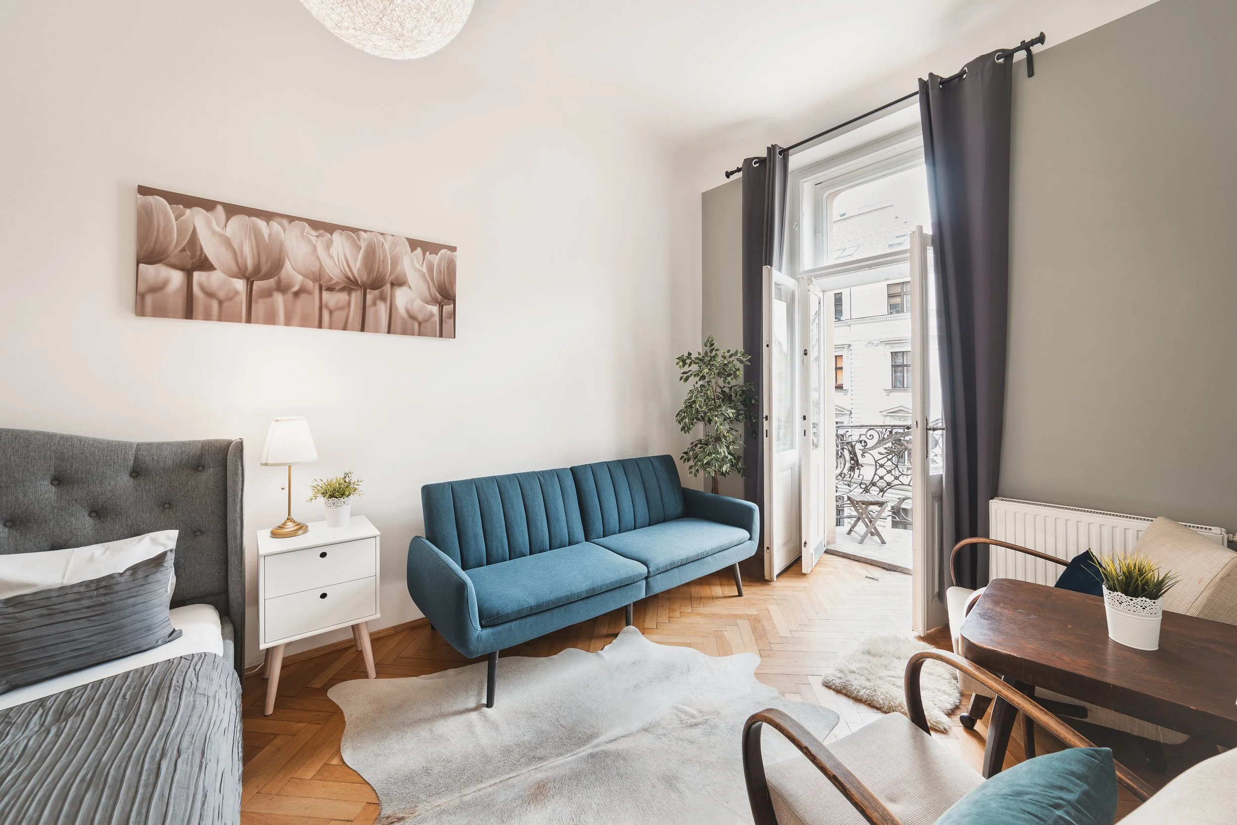 A bright living room with a bed, nightstand with lamp, a blue sofa, a small round table with a plant, a window with curtains, and balcony doors with a view of neighboring buildings.