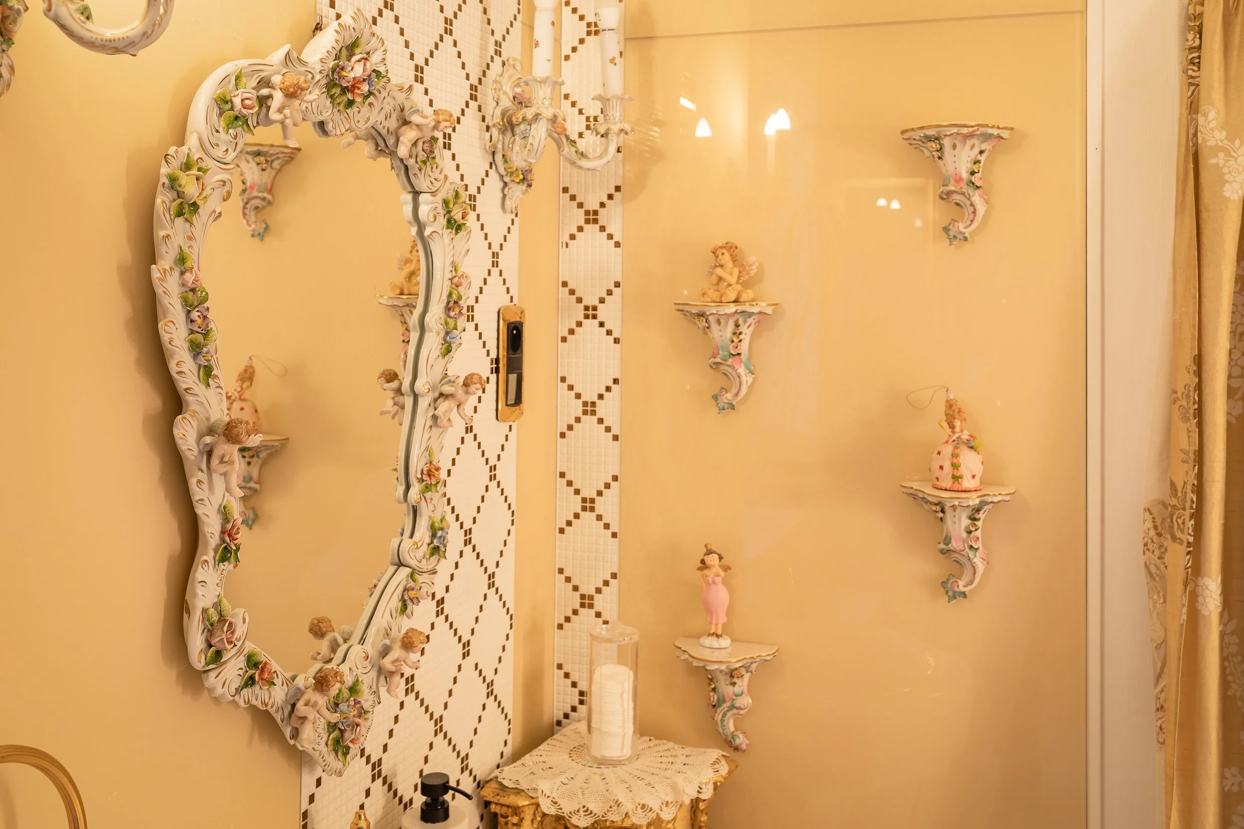 Decorative wall with floral and cherub-themed porcelain shelves and a mirror with floral details. Small cherub figurines are placed on the shelves, along with a candle holder and vases. The wall has a tiled pattern and a curtain is visible on the rig